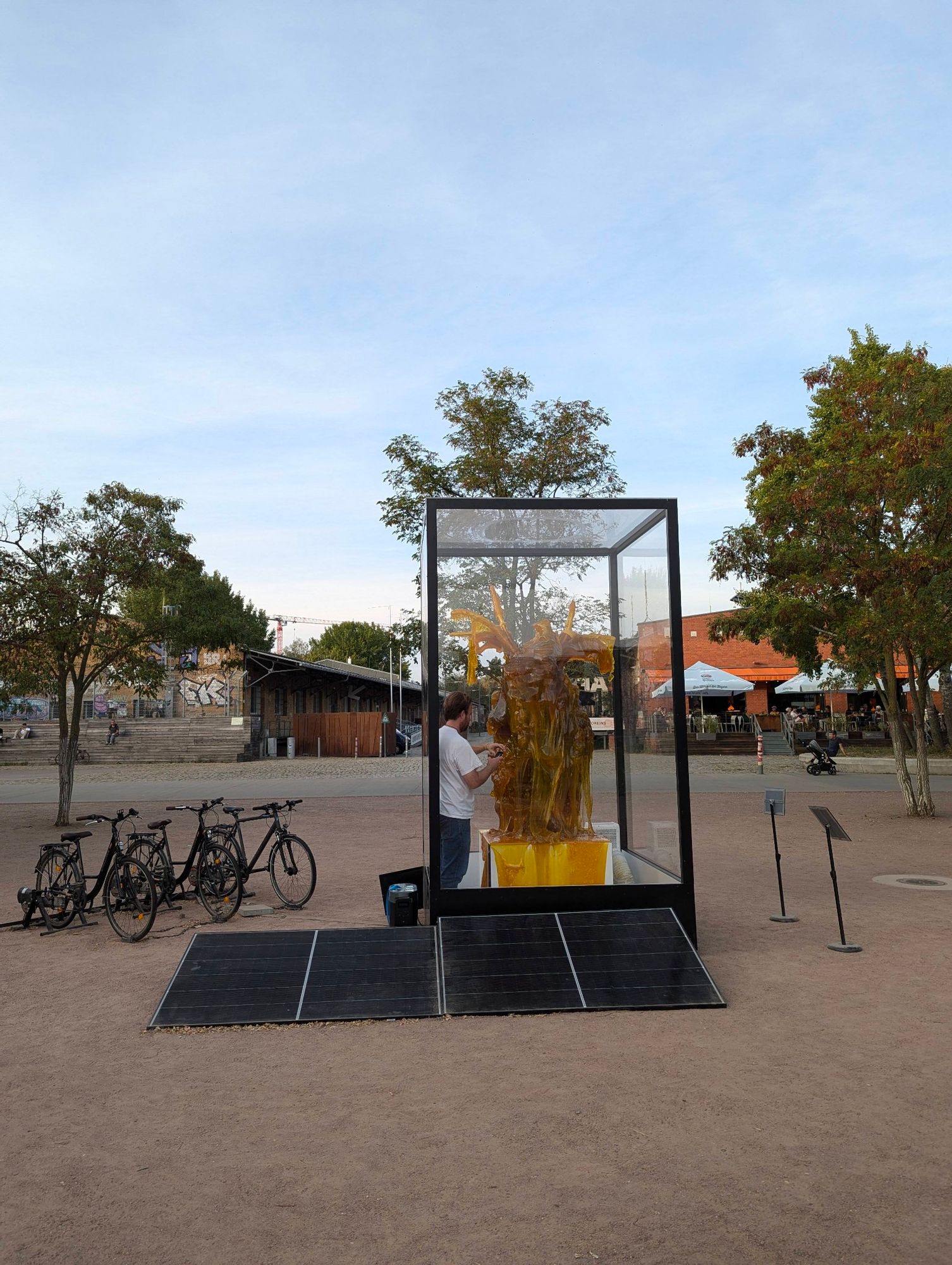 Eine Skulptur aus Zucker in Form eines menschlichen Herzens in einem Kasten mit Plexiglas umgeben. Die Skulptur ist teilweise geschmolzen durch die Hitze. 
Der Künstler, der das Herz erschaffen hat, ist gerade im Kasten und fotografiert von nahem den geschmolzenen Zucker. 
Links im Bild sieht man ein paar Fahrräder, welche einen Generator antreiben, der die Skulptur kühlen soll.