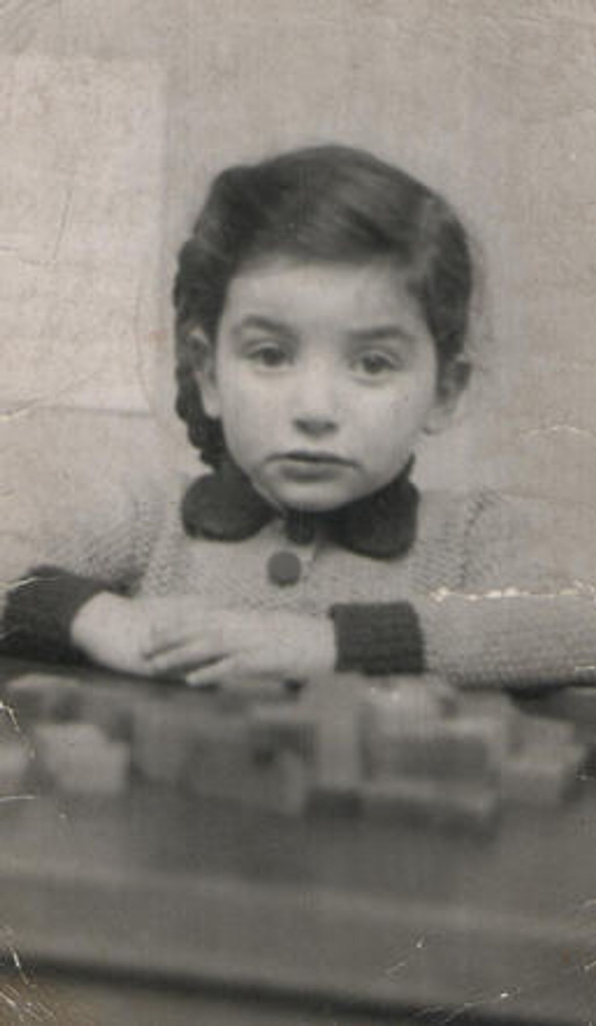 A vintage black and white photo of a young child seated at a table, wearing a sweater, with a thoughtful expression and hands clasped in front.