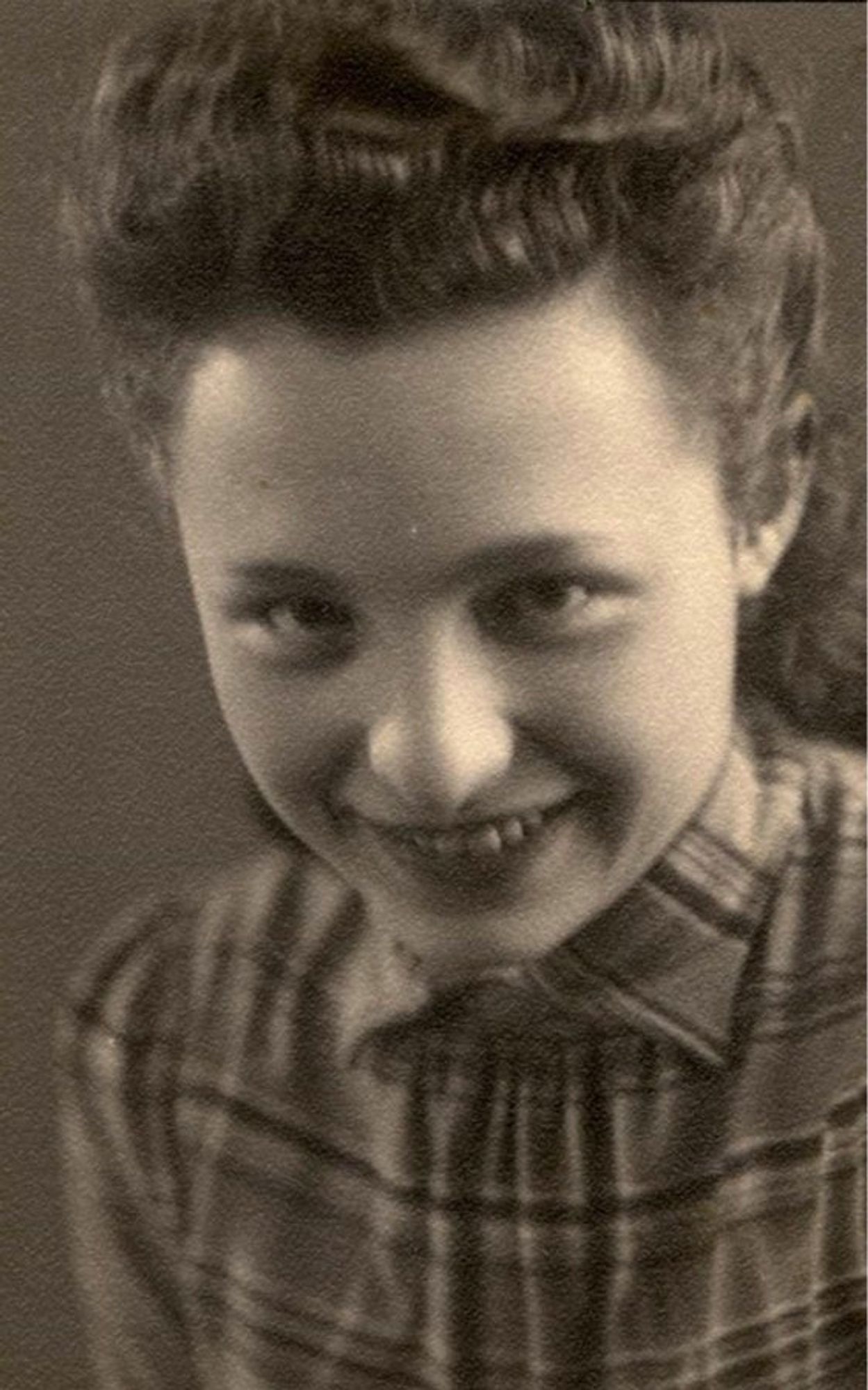 Portrait photo of a woman. She has long hair pinned back. Her fringe is attended in a wave. She is smiling - exposing her upper teeth. She is wearing a plaid blouse buttoned up to her neck.