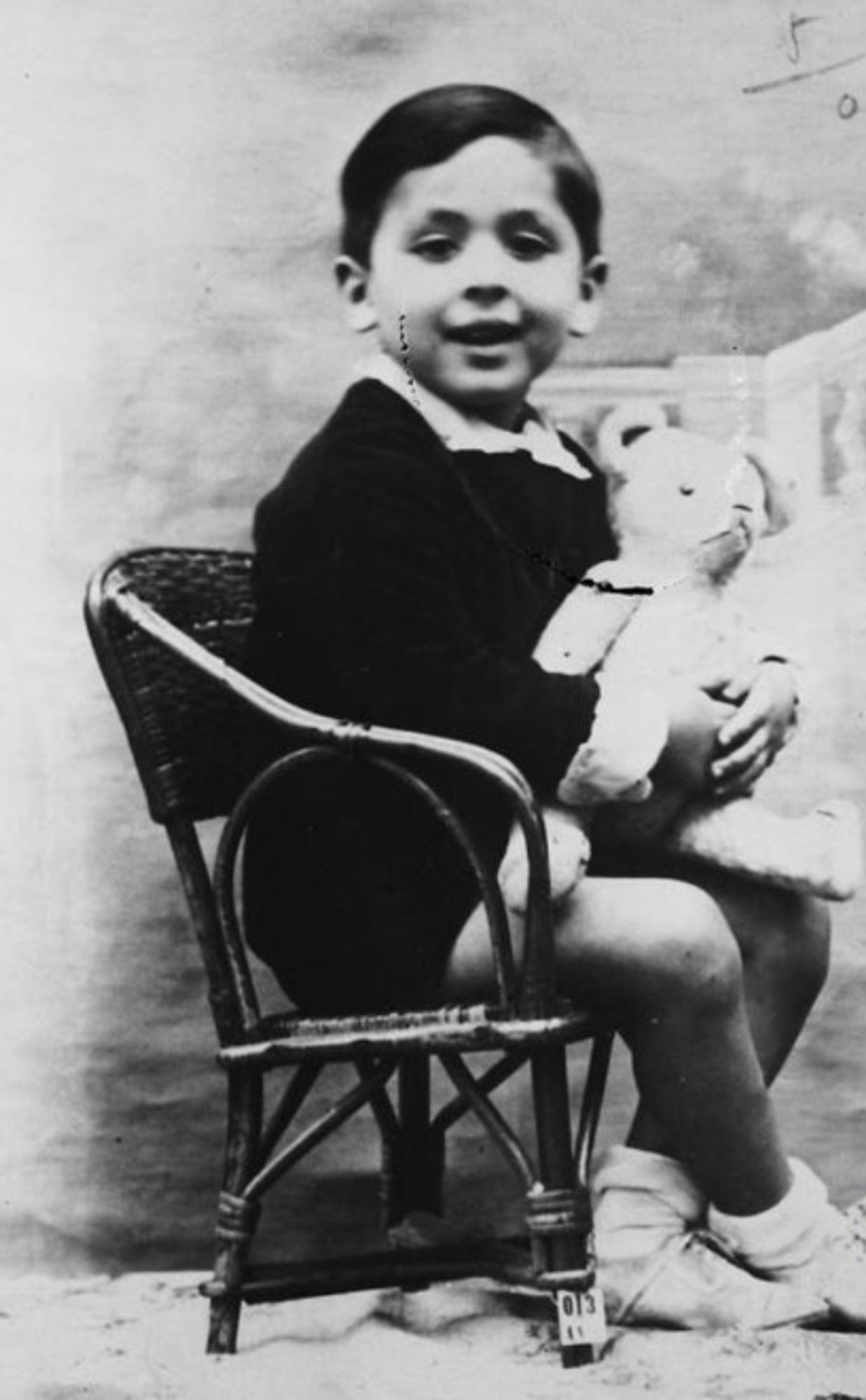 A vintage photo of a young boy seated on a chair, holding a teddy bear.