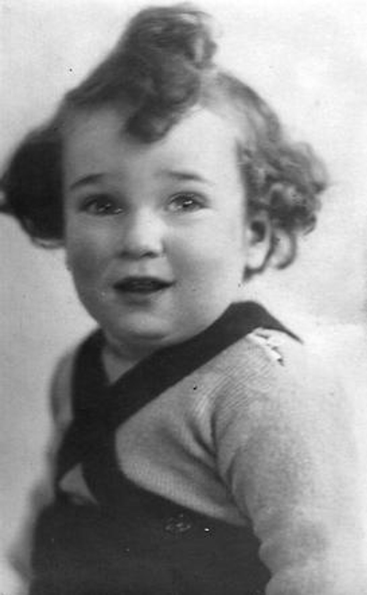 Vintage black and white photo of a young boy with curly hair, smiling and wearing a dark sweater with a shoulder strap.