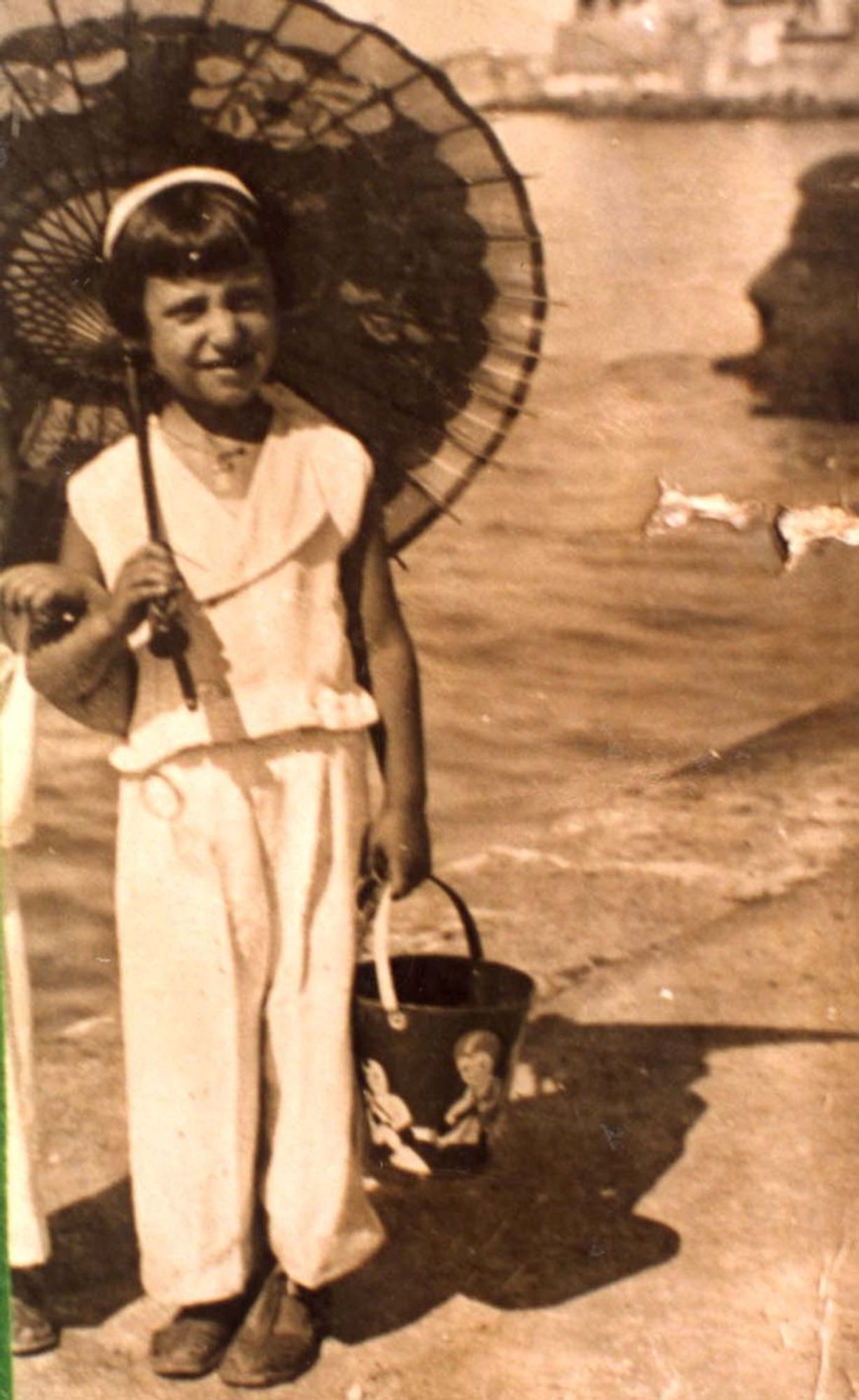 A vintage black and white photograph of a girl holding an umbrella and a bucket, dressed in a light-colored outfit, standing by a body of water.