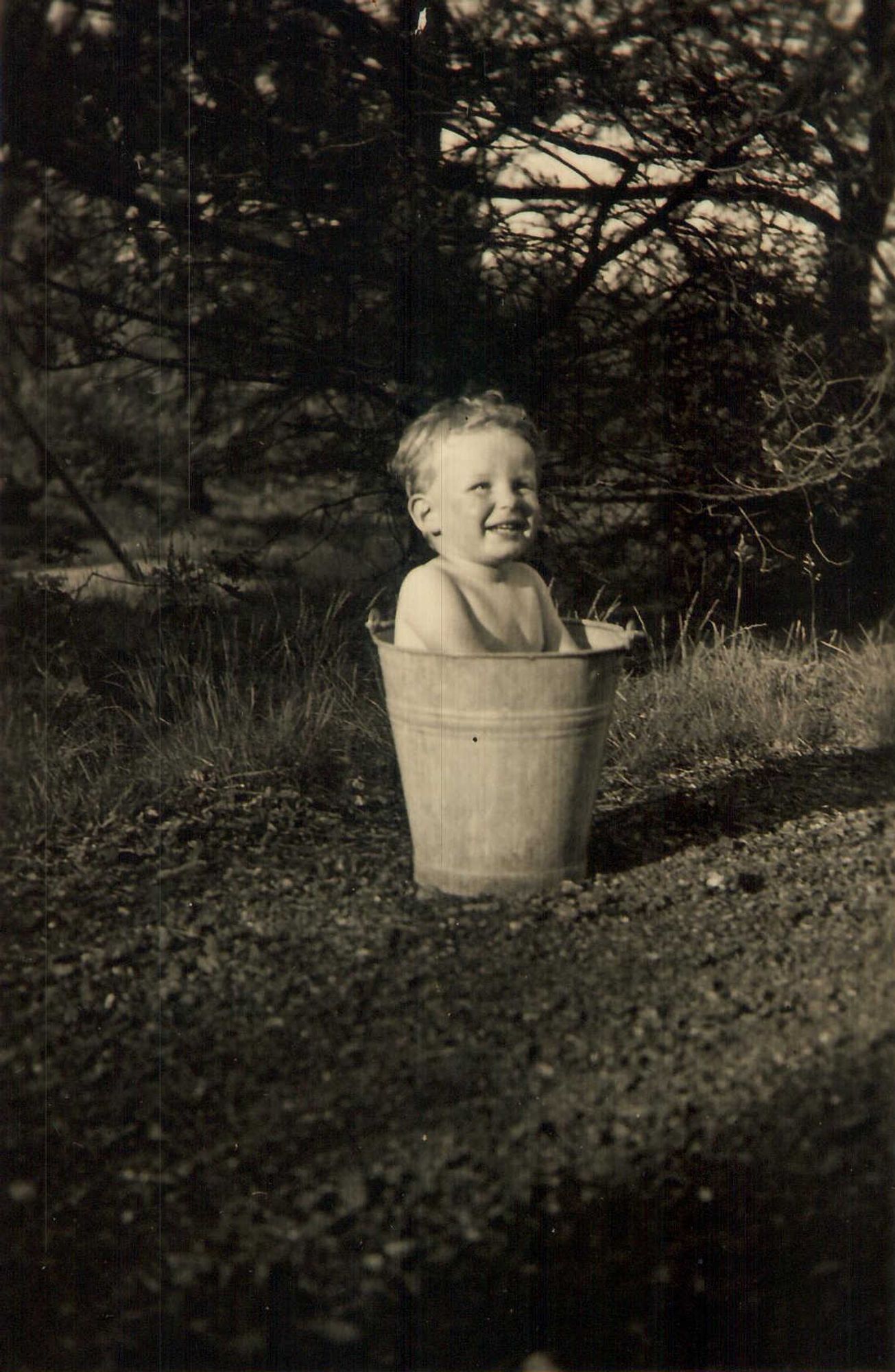 A sepia-toned photograph of a young child smiling joyfully while sitting inside a large metal bucket outdoors. The child appears to be enjoying a playful moment, with trees and grass surrounding them in a natural setting.