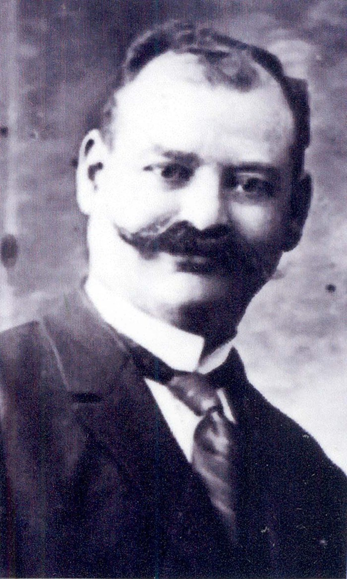 Portrait of a man, featuring a close-up of the individual with a prominent mustache, wearing a dark suit and tie.