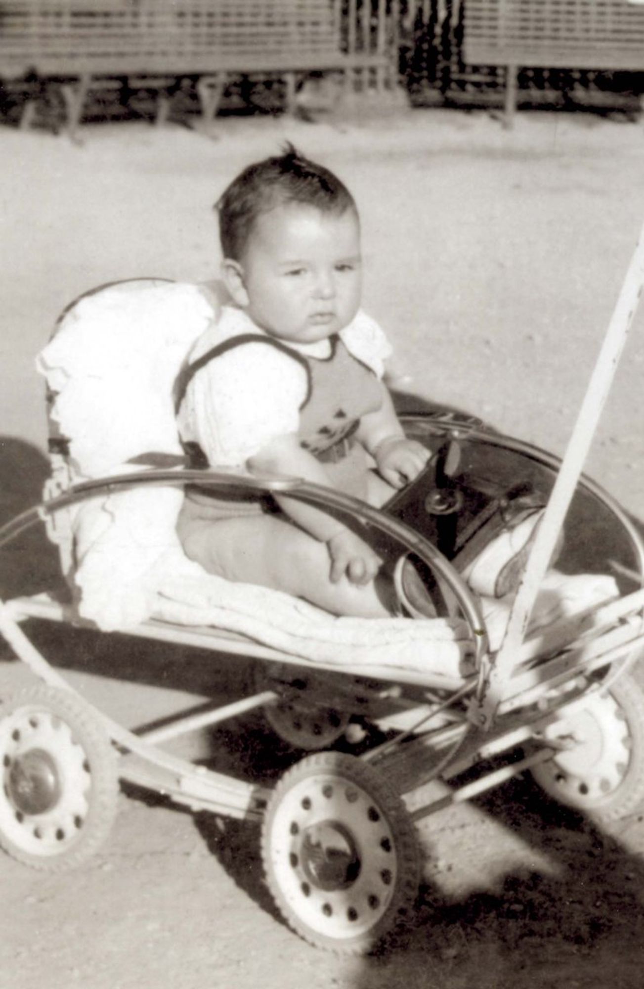 A vintage black and white photo of a baby sitting in a small baby stroller, looking directly at the camera. The setting appears to be outdoors with a fence visible in the background.