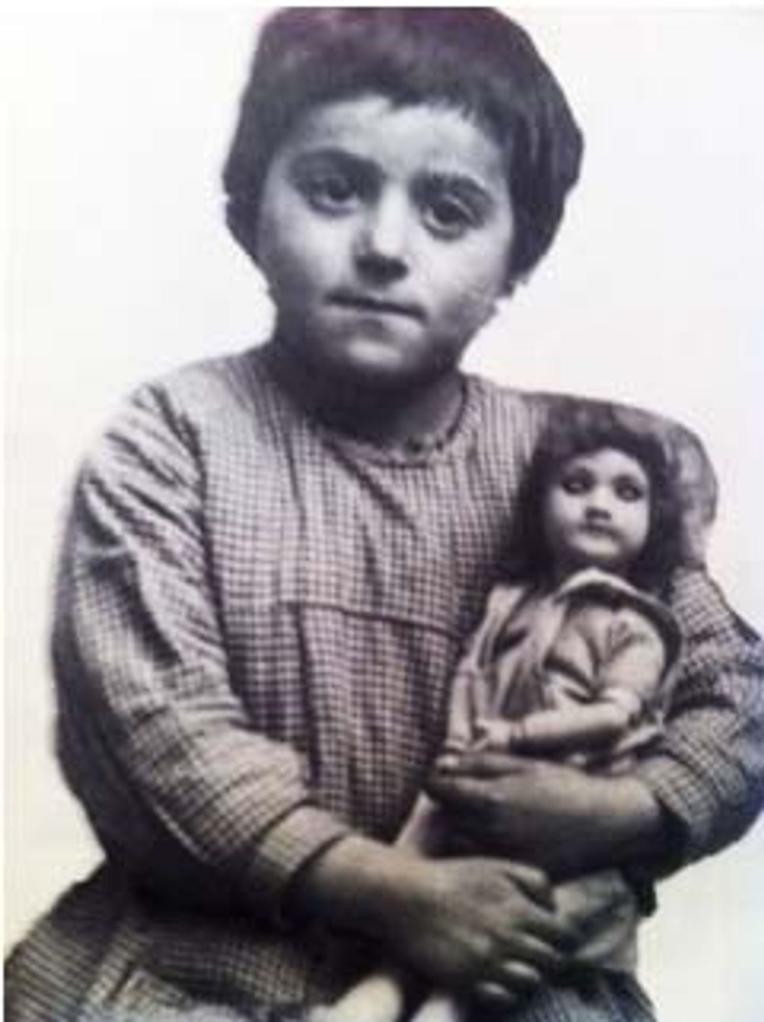 Vintage black and white photograph of a girl holding a doll, wearing a checkered outfit.
