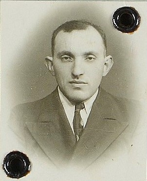 Vintage passport photograph of a man in a formal suit, with a serious expression. The photograph has two black binding holes on the upper edge.