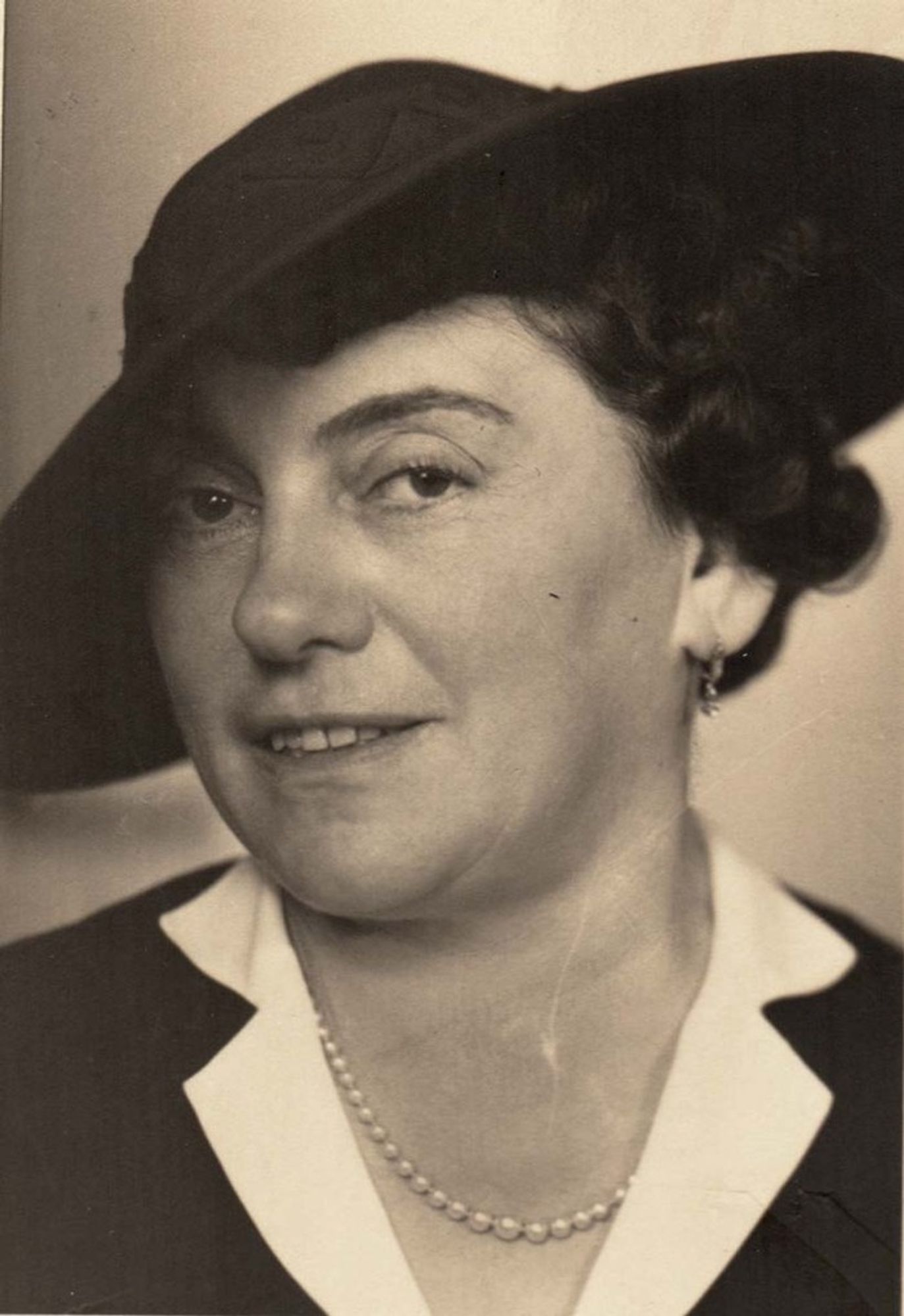 Vintage portrait of a woman wearing a dark hat and a light-colored blouse with a dark jacket, accessorized with a pearl necklace.