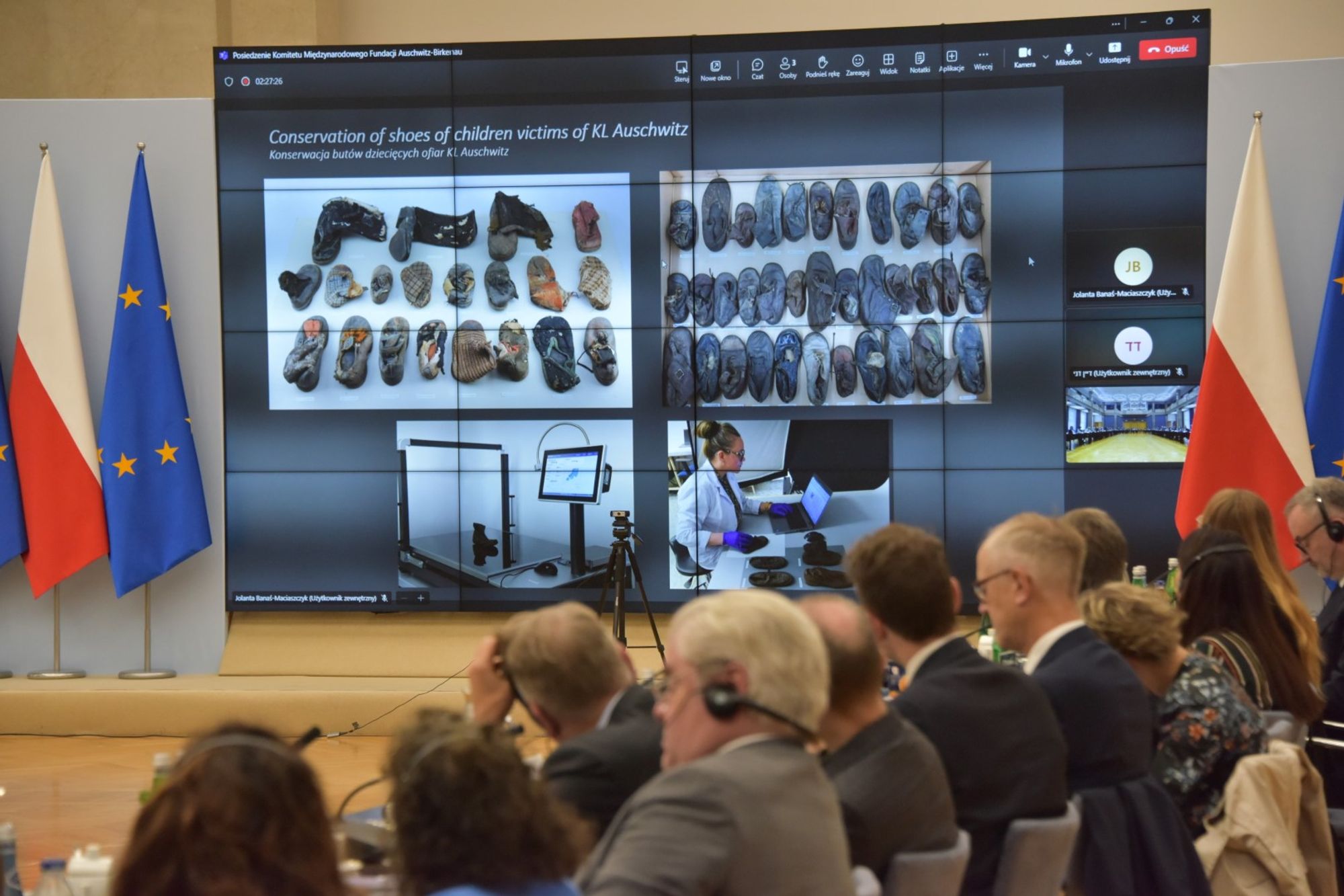 A large screen in a conference room displays images of children’s shoes being conserved as part of a presentation titled ‘Conservation of shoes of children victims of KL Auschwitz.’ The audience, including people in suits wearing headsets, sits in rows, with two flags — the European Union flag and the Polish flag — positioned on either side of the screen.