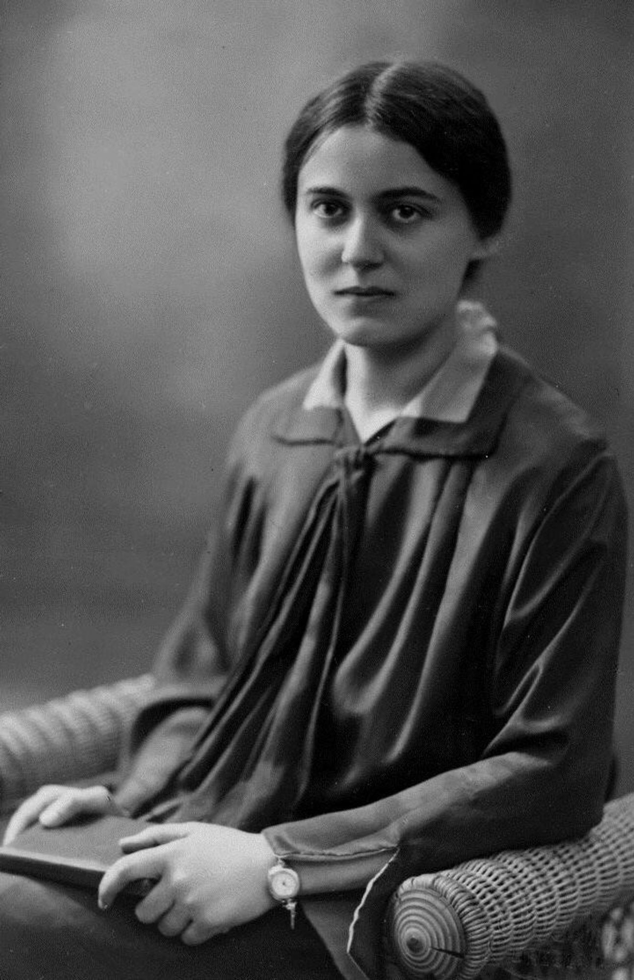 Black and white portrait of Edith Stein seated with hands clasped, wearing a dark blouse and a wristwatch, looking directly at the camera.