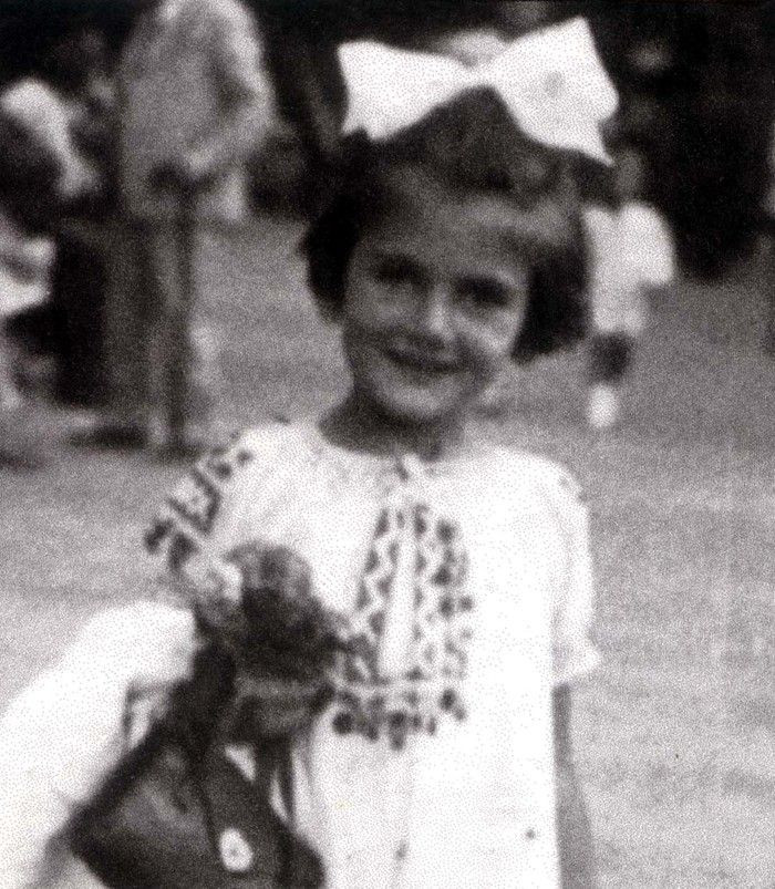 A vintage black and white photo of a young girl smiling at the camera. The child is wearing a light-toned dress with a decorative pattern and a large bow in their hair. The background is blurred with indistinct figures.