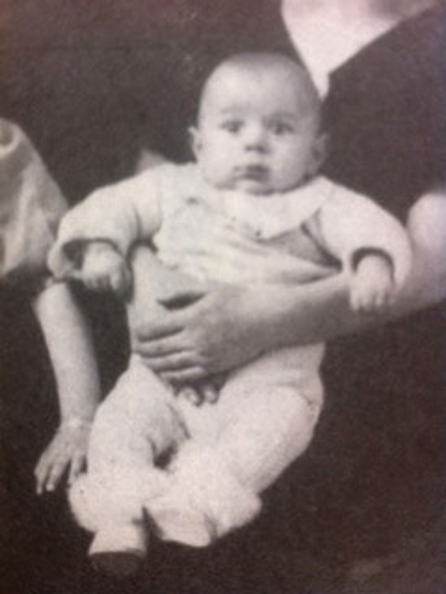 Black and white historical photo showing a baby being held by an adult, only the adult's arms and torso are visible.