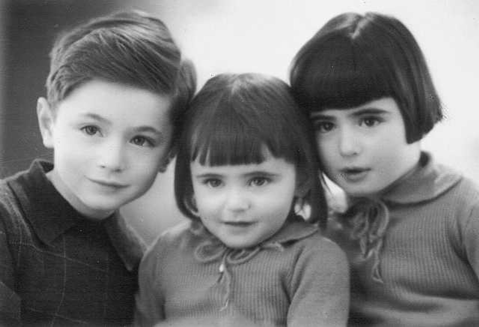 Three young children posing closely for a black and white photo. The boy on the left has styled hair and wears a sweater, the middle child has short hair and a turtleneck, and the girl on the right has a bob haircut and also wears a sweater.