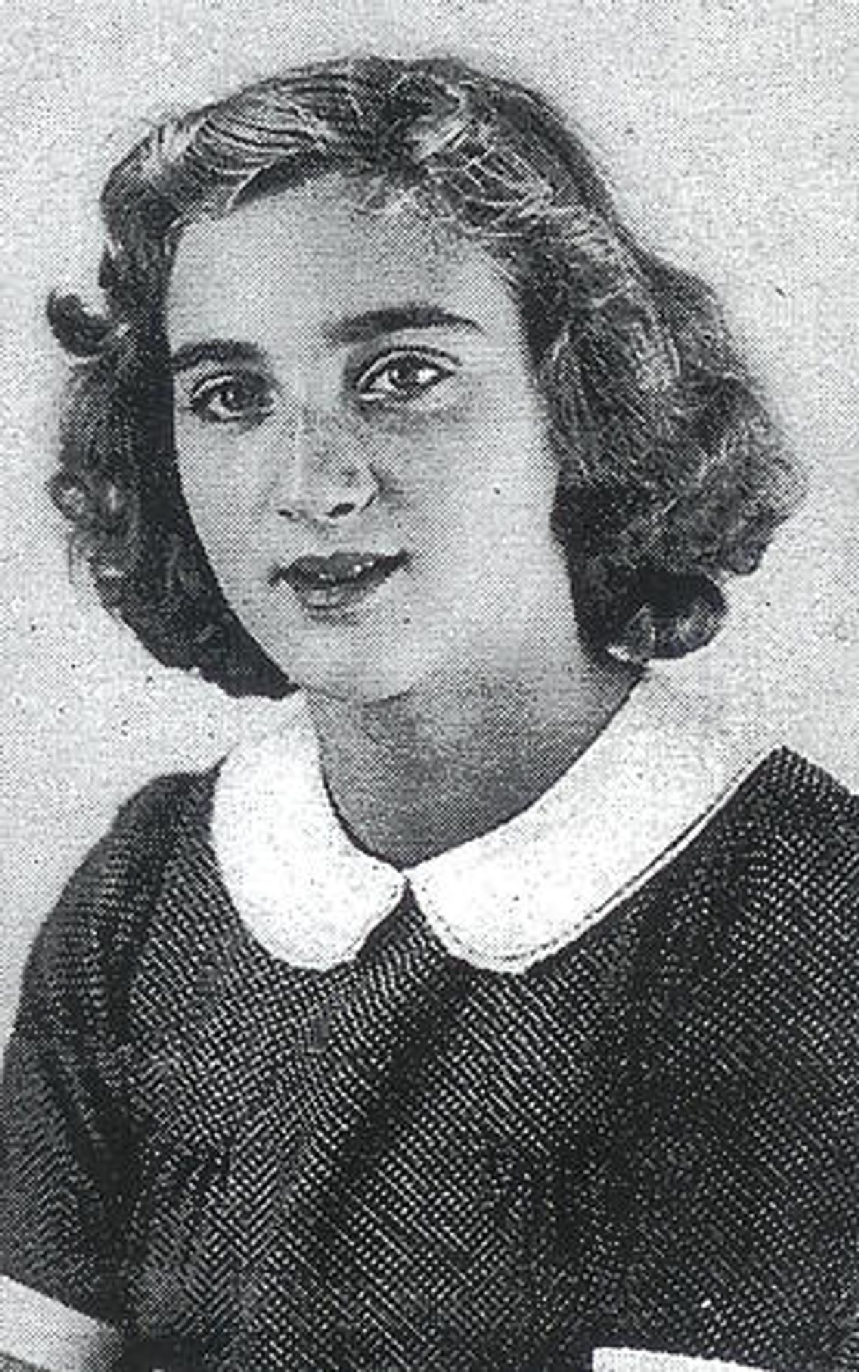 Portrait photograph of a teenage girl carded above the waist. She has long wavy hair reaching her neck and a slightly open mouth. She is wearing a fine dark checked blouse with short sleeves and a wide white collar. 