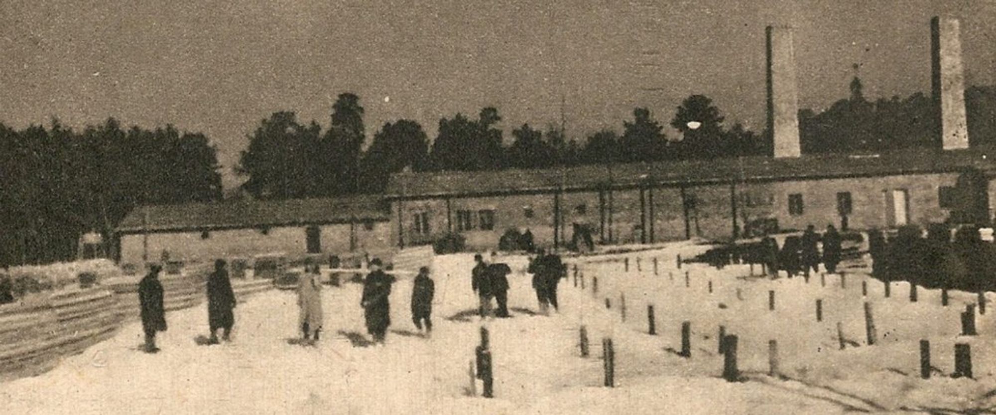 Black and white historical photograph of a group of people working in a snowy field , with two chimneys and a long building in the background. The building was gas chamber and crematorium IV at Auschwitz II-Birkenau camp.