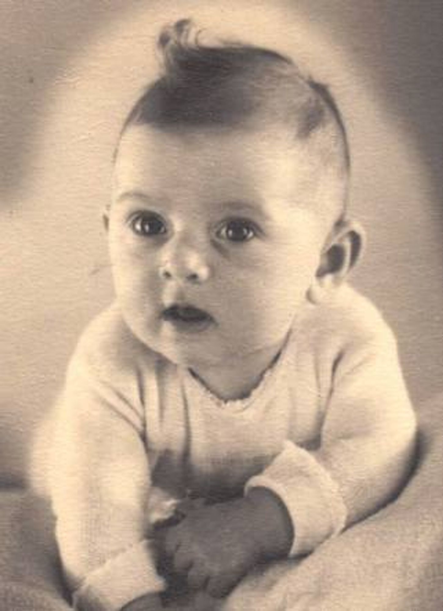 A little girl with a curl on her head lies on a blanket. Photographed from the front. She is leaning on her arms and raising her head. Her mouth is open and her eyes are wide open.