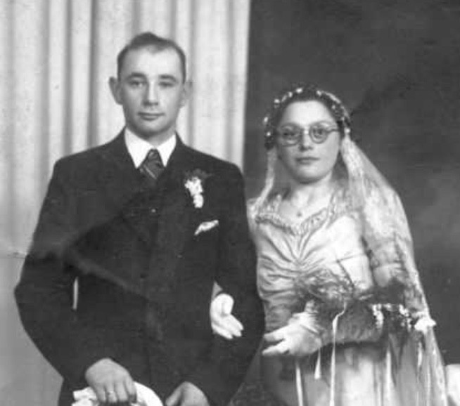 Vintage wedding photo of a couple, featuring a man in a dark suit and another in a detailed bridal gown and veil, in a formal studio setting.