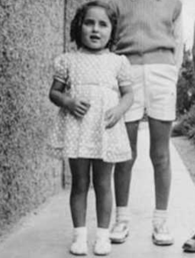 Black and white photo of a girl in a polka dot dress standing next to an adult, only the lower body of the adult is visible. They are both on a narrow pathway with a wall on one side.