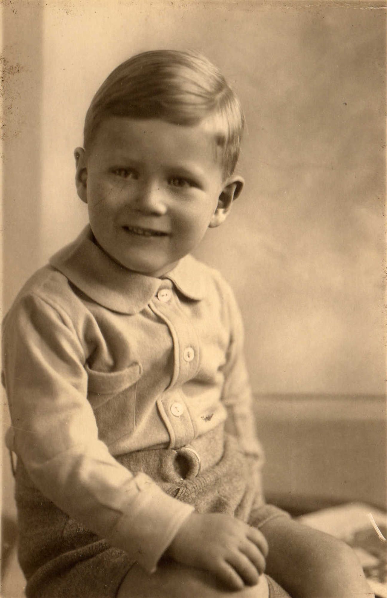 A vintage sepia-toned portrait of a young boy with short hair, smiling and sitting with one arm resting on their knee.