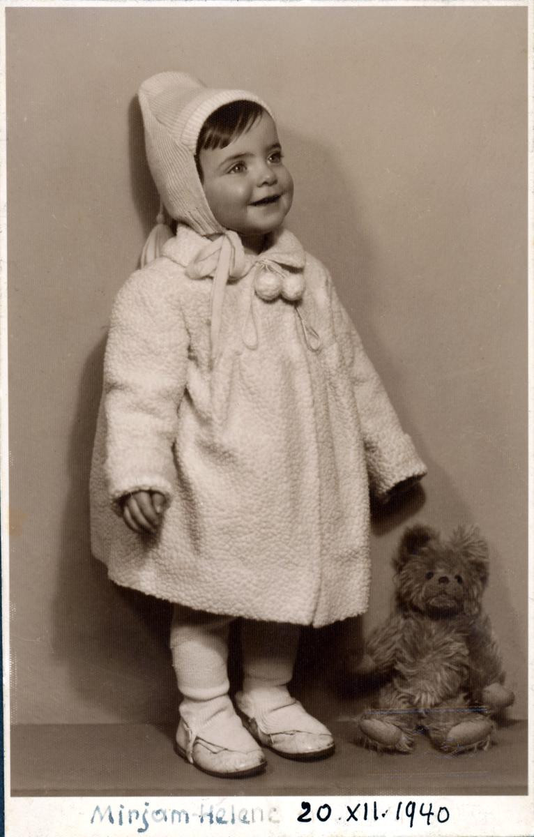 A vintage black and white photo showing a smiling girl in a hooded coat and white shoes standing next to a plush teddy bear.