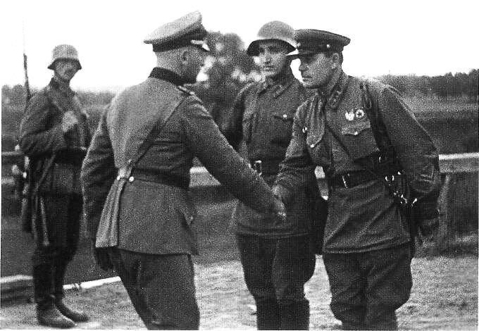 Black and white historical photo of four uniformed soldiers - from Nazi German and Soviet Union standing outdoors. One officer is shaking hands with another while two are observes.