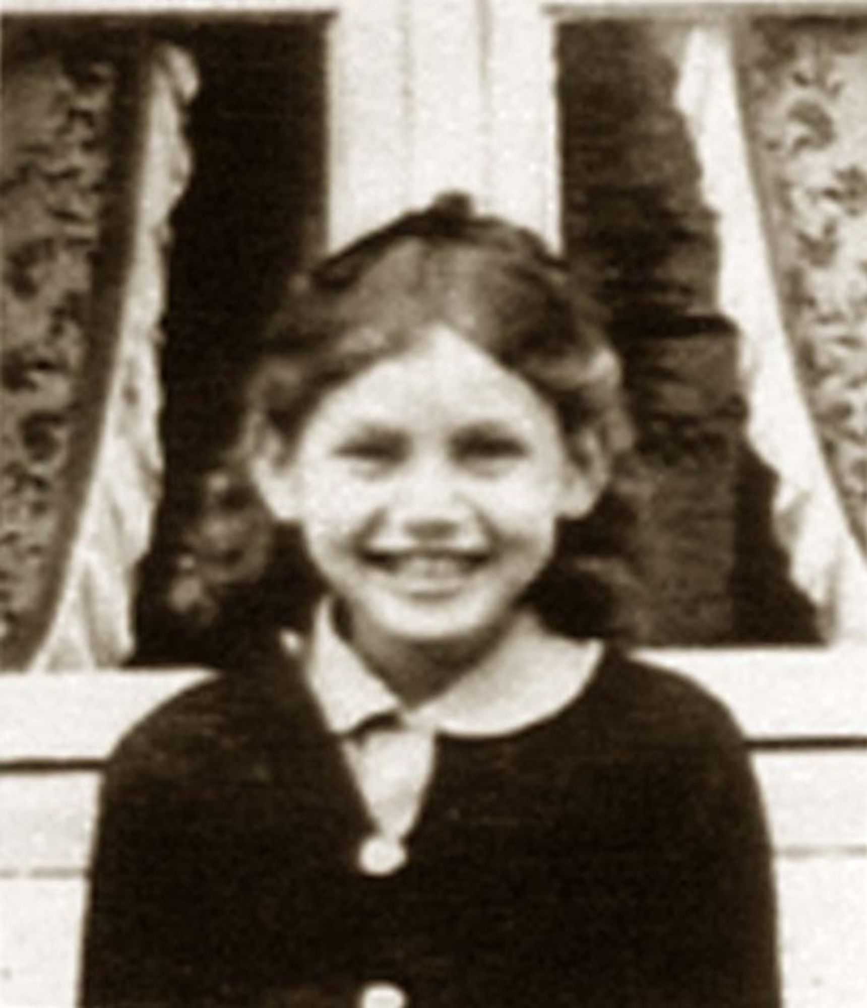 A vintage black and white photo of a young girl smiling in front of a curtained window.