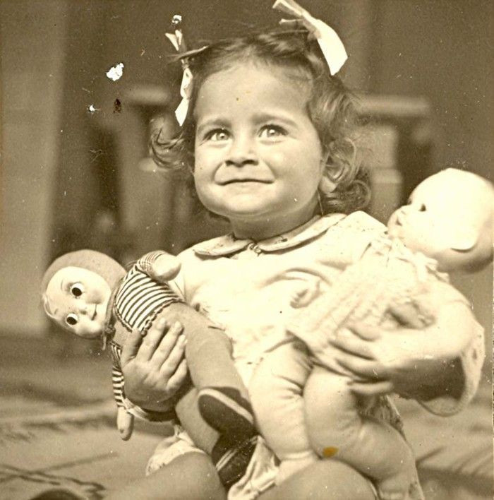 A vintage photograph of a smiling girl holding two dolls.