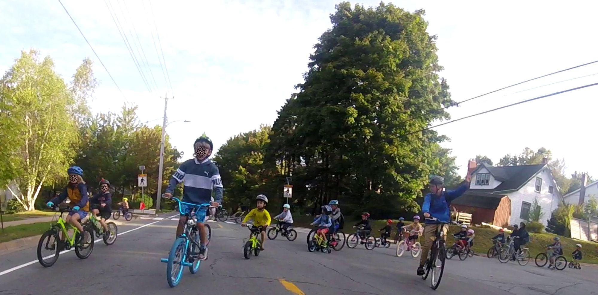 20 kids on bikes and scooters filter onto a road in Dartmouth, NS on their way to school, accompanied by some parents.