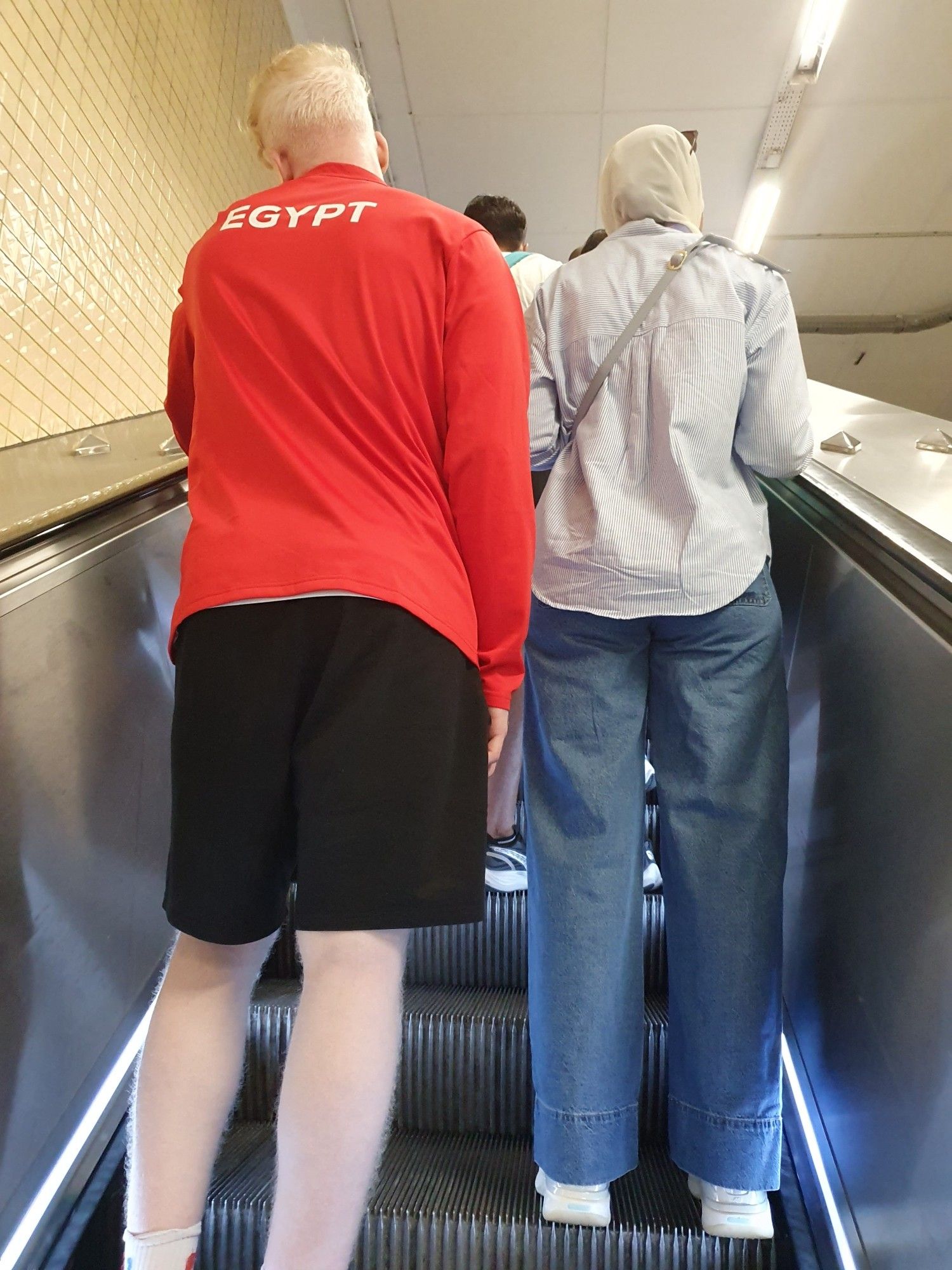 On a métro escalator going up, several men seen from behind, one of them wearing a red jacket marked "Egypt" over sportswear.