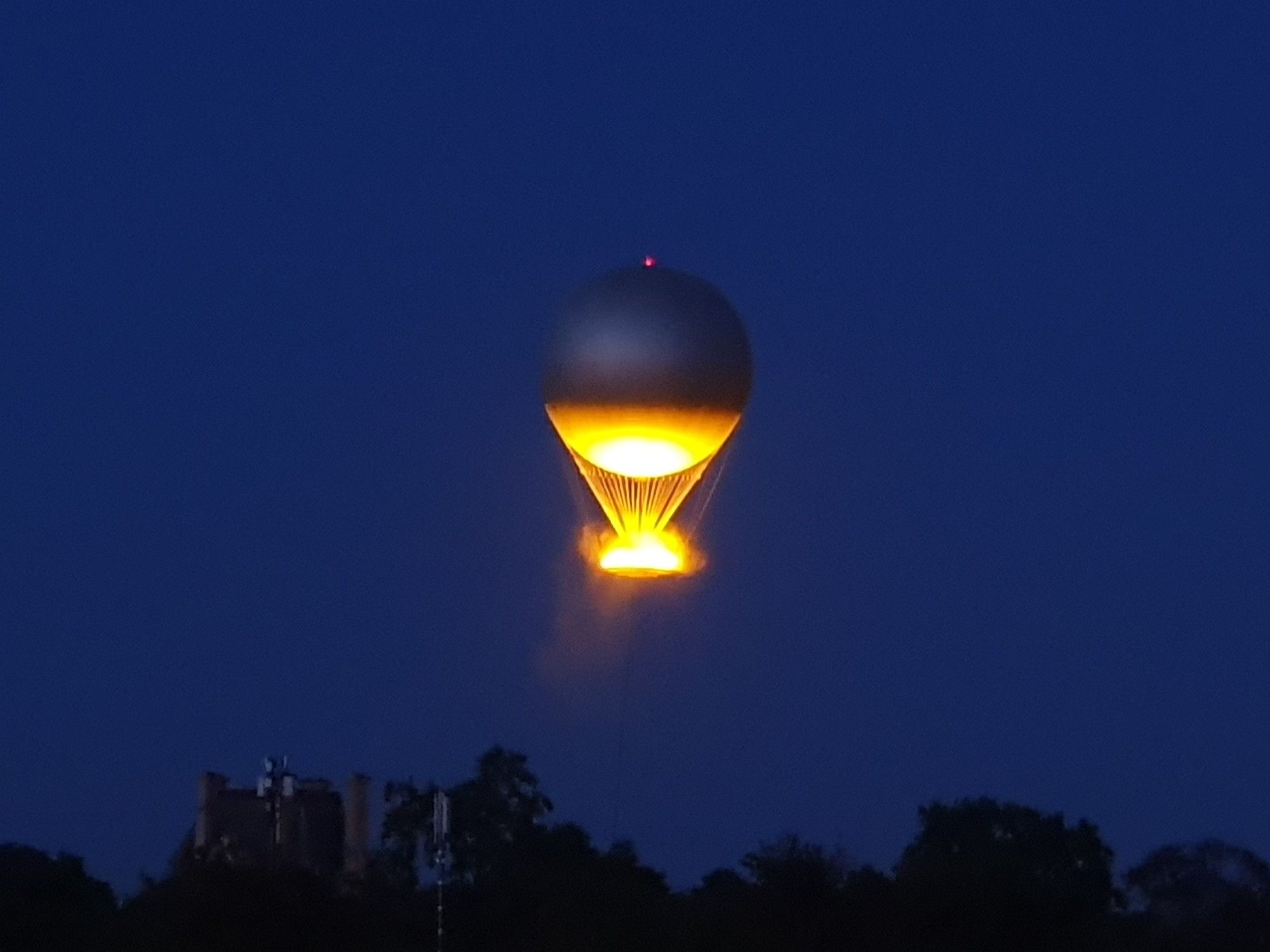 A tethered hot-air balloon in the night sky, below a faint dark silhouette of trees and buildings. A flame burns in the flat basket below the round balloon, so that the basket and the underside of the balloon are yellow