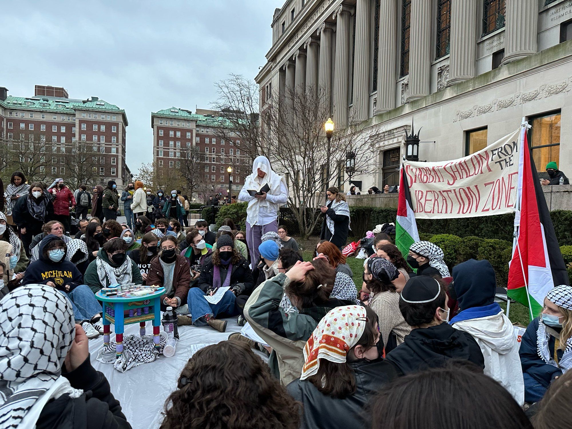 A group of students, some wearing yarmulkes, many wearing keffiyehs, sit gathered in Shabbat service on the lawn in front of Butler Library at Columbia University. A banner flanked by flags of Palestine reads SHABBAT SHALOM FROM THE LIBERATED ZONE