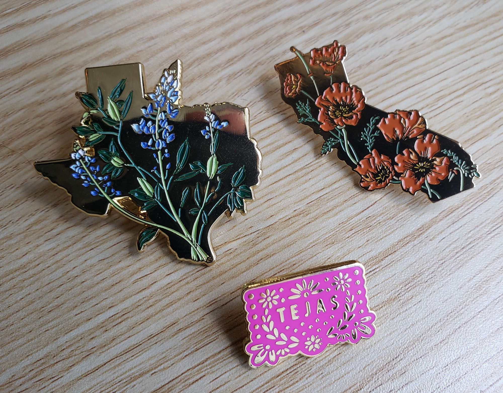 enable pins featuring a brass silhouette of texas with bluebonnets, a brass silhouette of california with poppies, and a pink papel picado that says tejas.