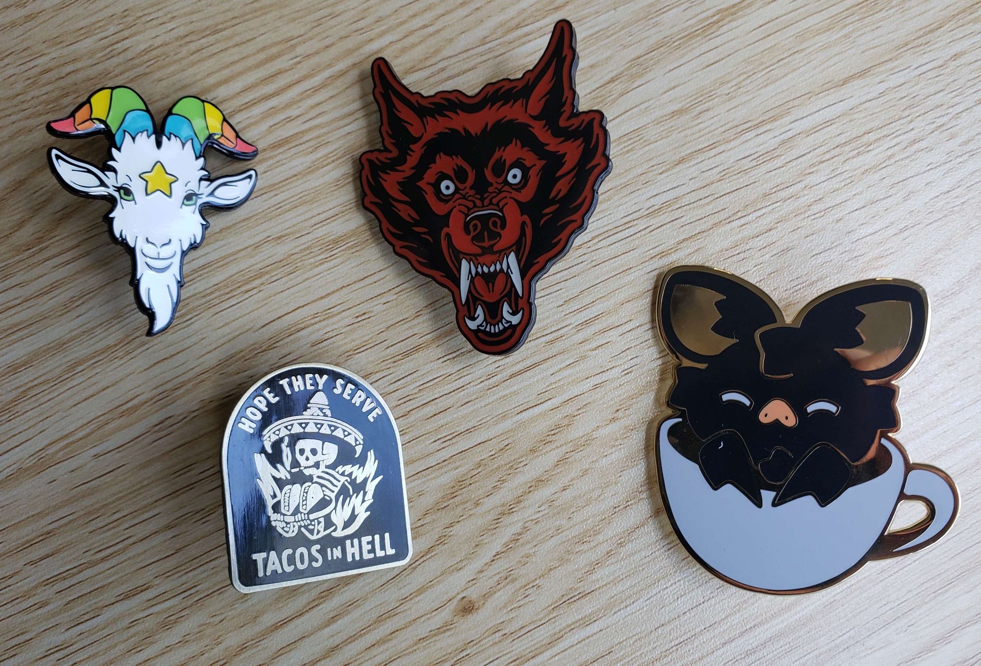 enamel pins that feature a white goat with rainbow horns, a red and black snarling werewolf, a skeleton in a sombrero that says "i hope they serve tacos in hell", and a cute black bat in a coffee cup.