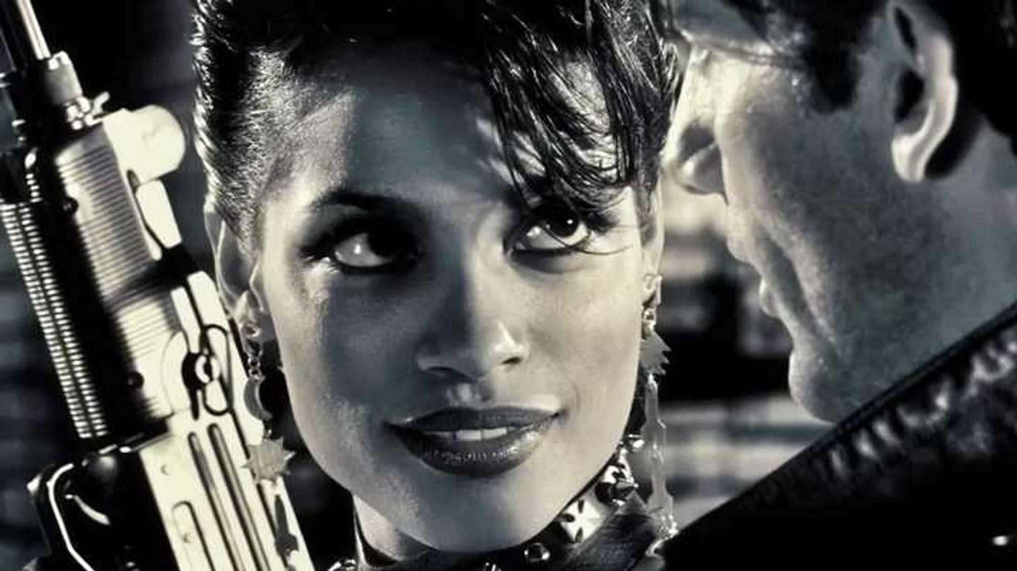 Rosario Dawson in the movie Sin City* as Gail (one of the Girls of Old Town**. Prostitute & enforcer***). Looking with desire, a little madness, a sexy smile and look. With kinky short hair and a leather necklace with metal crosses on it. Beautiful sexy smiling face. While holding an automated firearm showing her power as why she is an enforcer.

*Sin City: In the comic and movie Sin City is a Black & White Stylized, hard-boiled noir story set in the titular hellhole, which is 
overrun by corruption and decay.
**Old Town is a section of Sin City run by the local prostitute community, known as the Girls of Old Town.
***Enforcers are the armed, strong, powerful, lethal and not to forget the sexy protectors of the Old Town Girls. Trusting no man to do that for them.
Bonus: several quotes are world famous like “a dame to kill for” from this story.