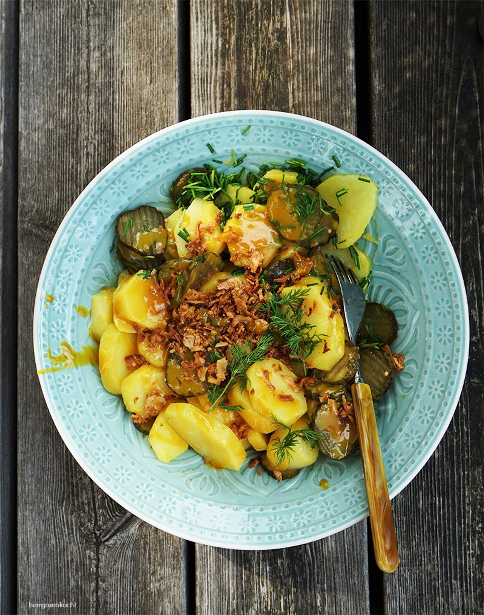 Grün-Grauer Holzuntergrund. Darauf ein hellgrüner runder, tiefer Teller mit Kartoffelsalat, Gurkenscheiben, gerösteten Zwiebeln und eine Senf-Sauce.