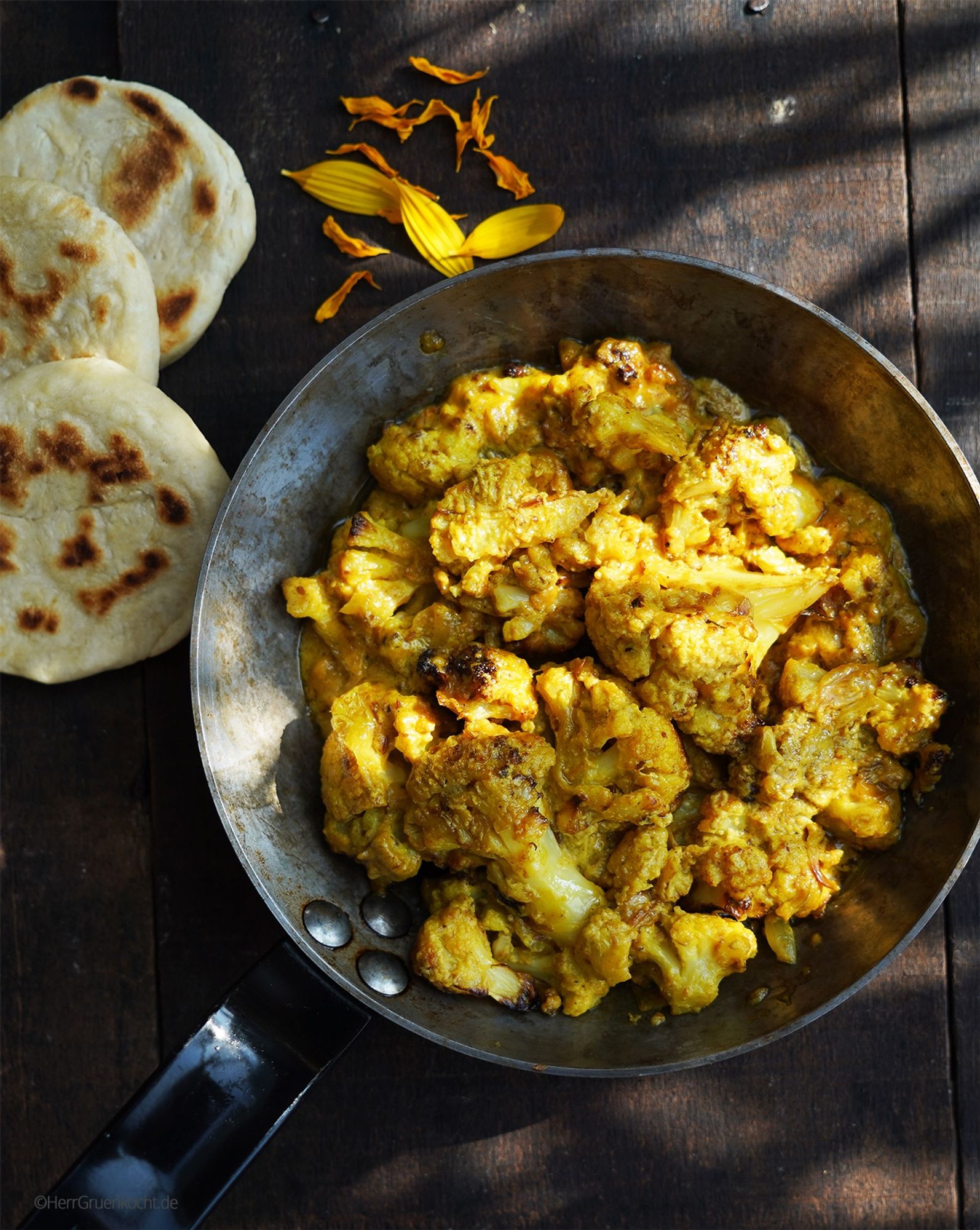 Dunkler Holzuntergrund. In der Mitte eine kleine, silberne Bratpfanne. Darin Gelbes Blumenkohlcurry. Neben der Pfanne: Kleine in einer Pfanne gebackene Joghurt-Küchlein mit Brandflecken.