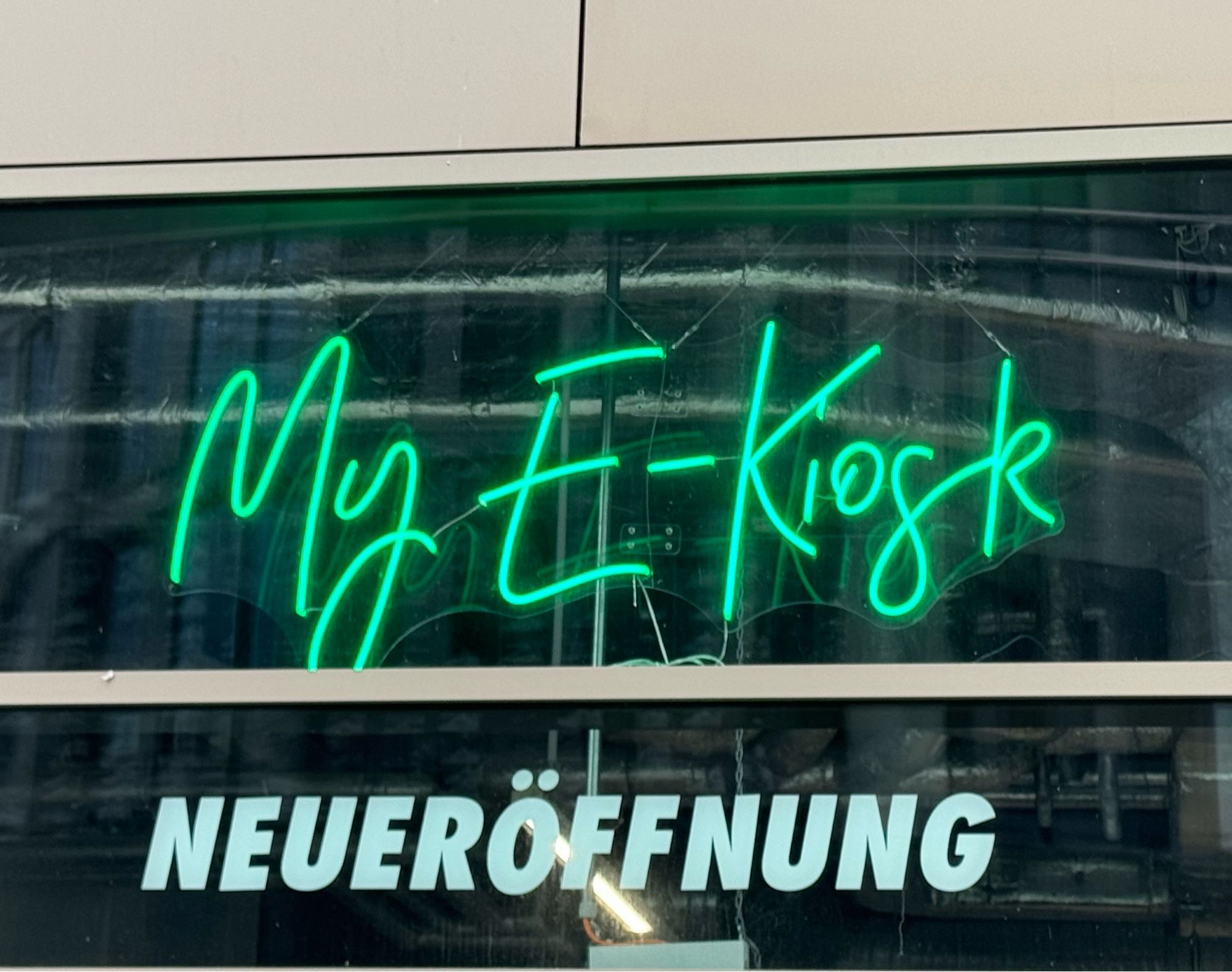 an einem Geschäft ist eine grüne Neon-Reklame angebracht ‚My E-Kiosk‘ darunter ‚Neueröffnung‘