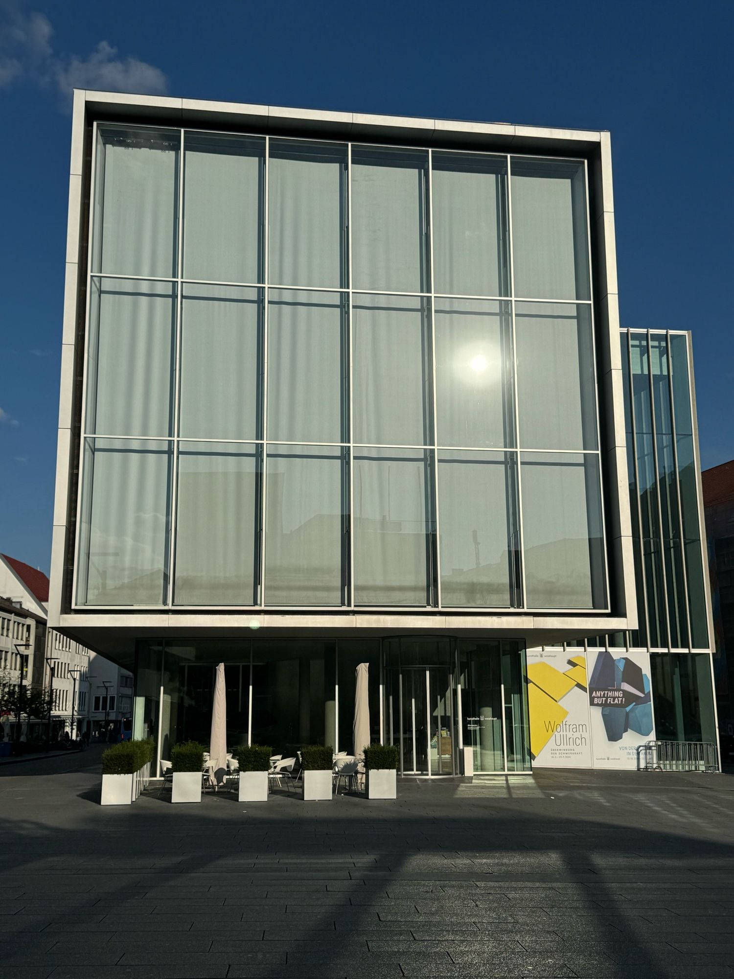 auf einem Platz steht ein Museum mit einer Glas- und Stahlfassade - die Scheiben sin senkrechte Fenster - in einem Fenster rechts spiegelt sich schwach die Sonne - vor dem Gebäude stehen fünf kleine Betonsockel