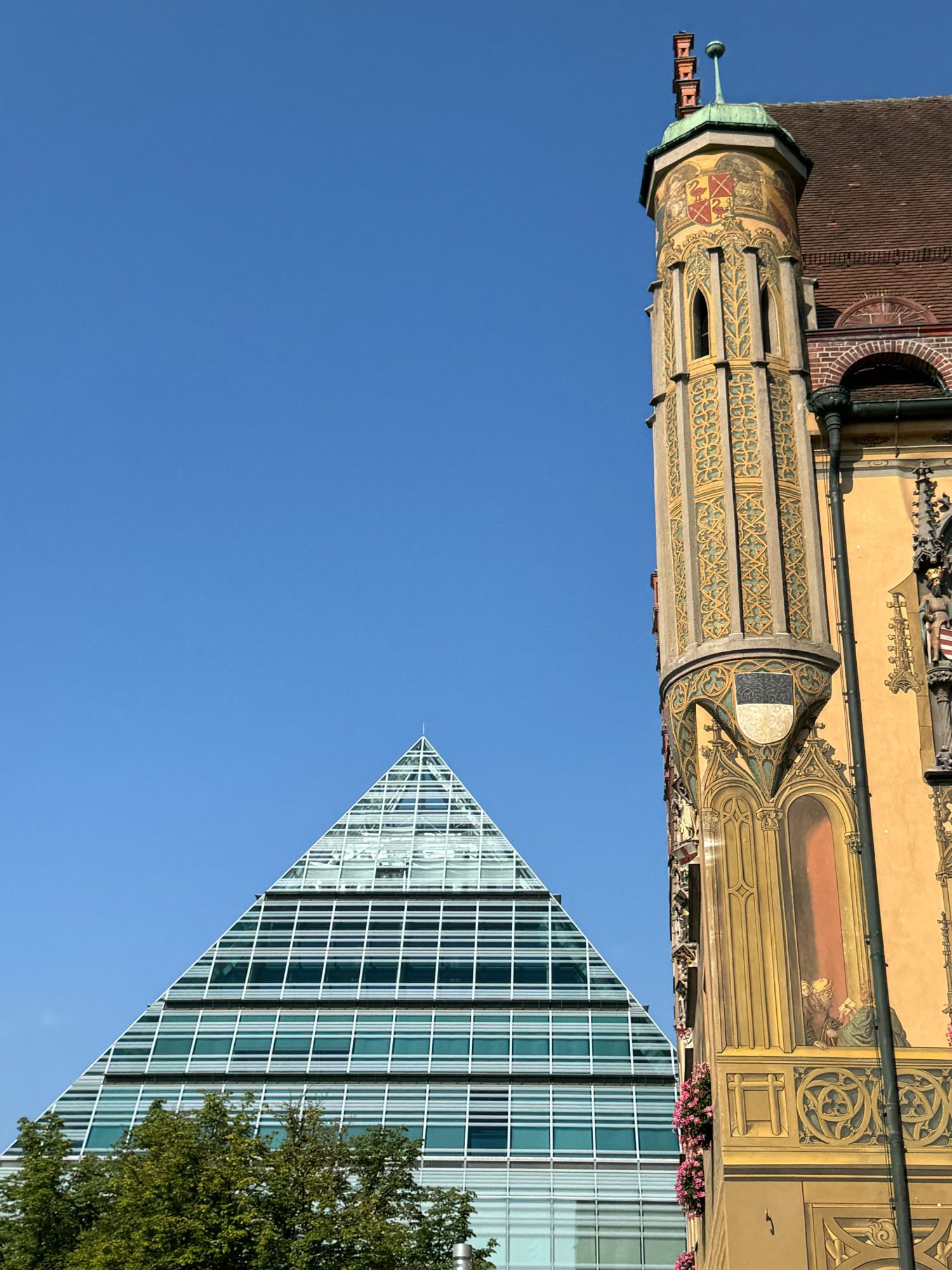 linker Bereich ein Gebäude in Form einer Pyramide aus Glas und Stahl - davor ein Baum - rechts ein Teil der Fassade vom historischen Rathaus mit Erker und bunter Bemalung - blauer wolkenloser Himmel