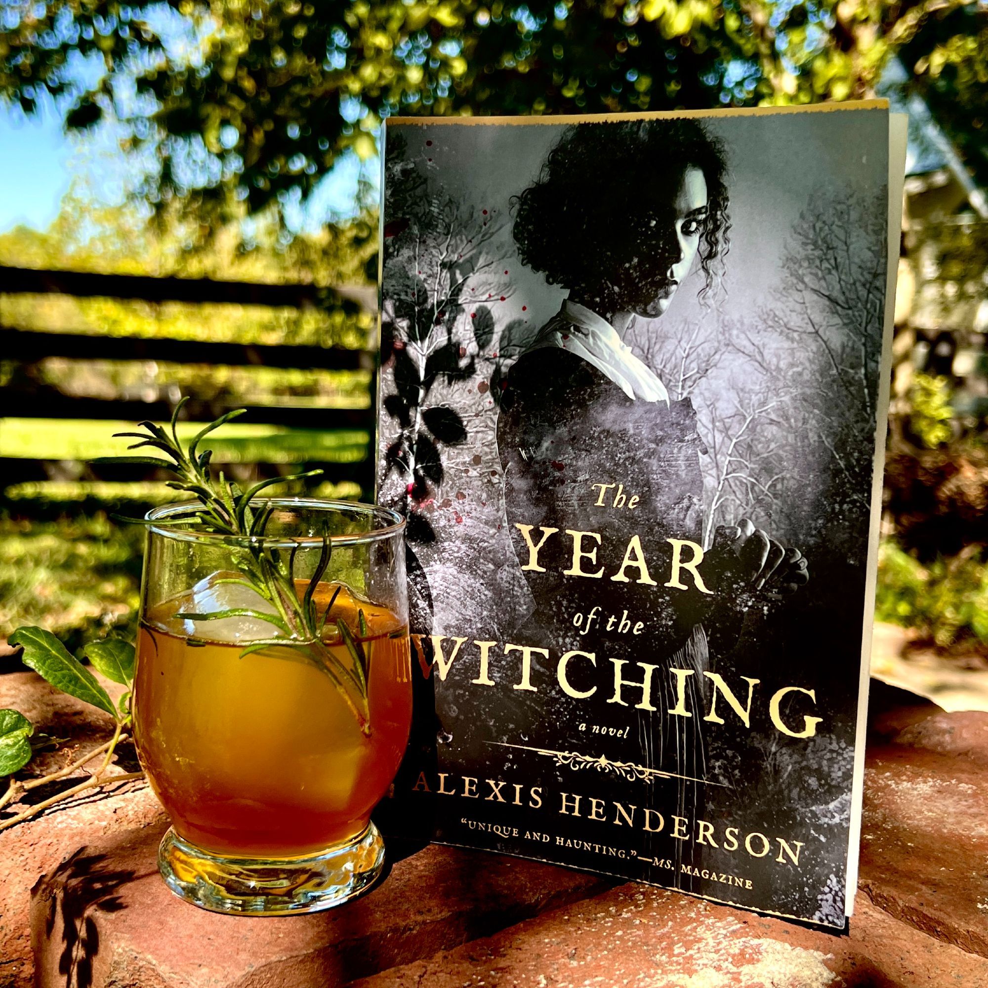 [Image desc: an old fashioned glass with a rich amber mocktail garnished with a sprig of rosemary sits amid autumn sun on a pile of bricks alongside a paperback copy of THE YEAR OF THE WITCHING by Alexis Henderson]