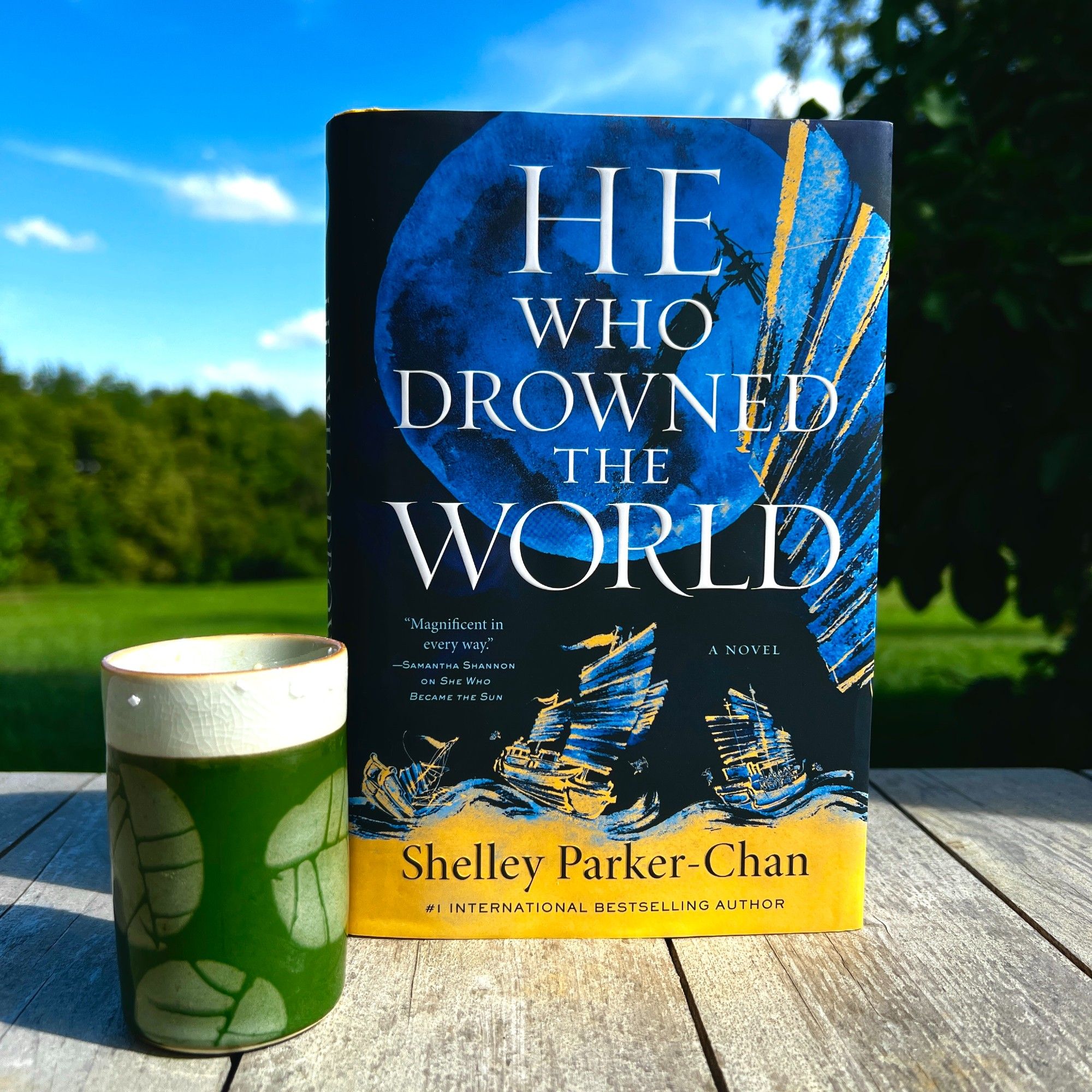 A copy of HE WHO DROWNED THE WORLD by Shelley Parker-Chan sits on a wooden surface against a blue sky and green fields. Beside the book is a green cylindrical tea cup with a white border rimmed with kosher salt.