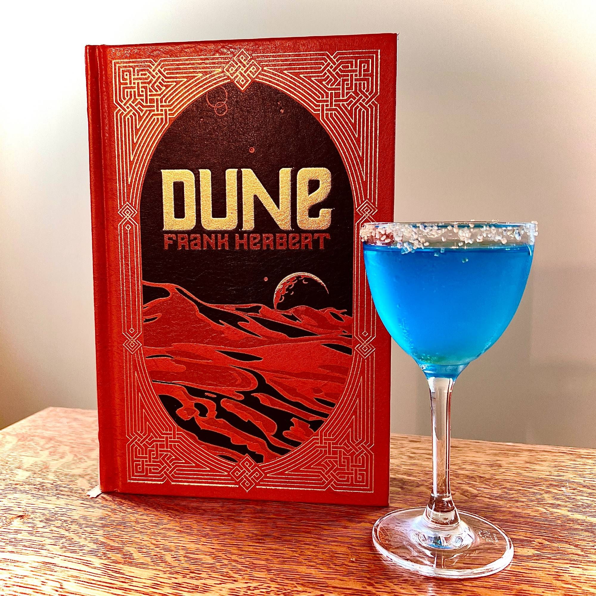 Dune by Frank Herbert alongside a bright blue cocktail in a stemmed glass rimmed with golden sugar.