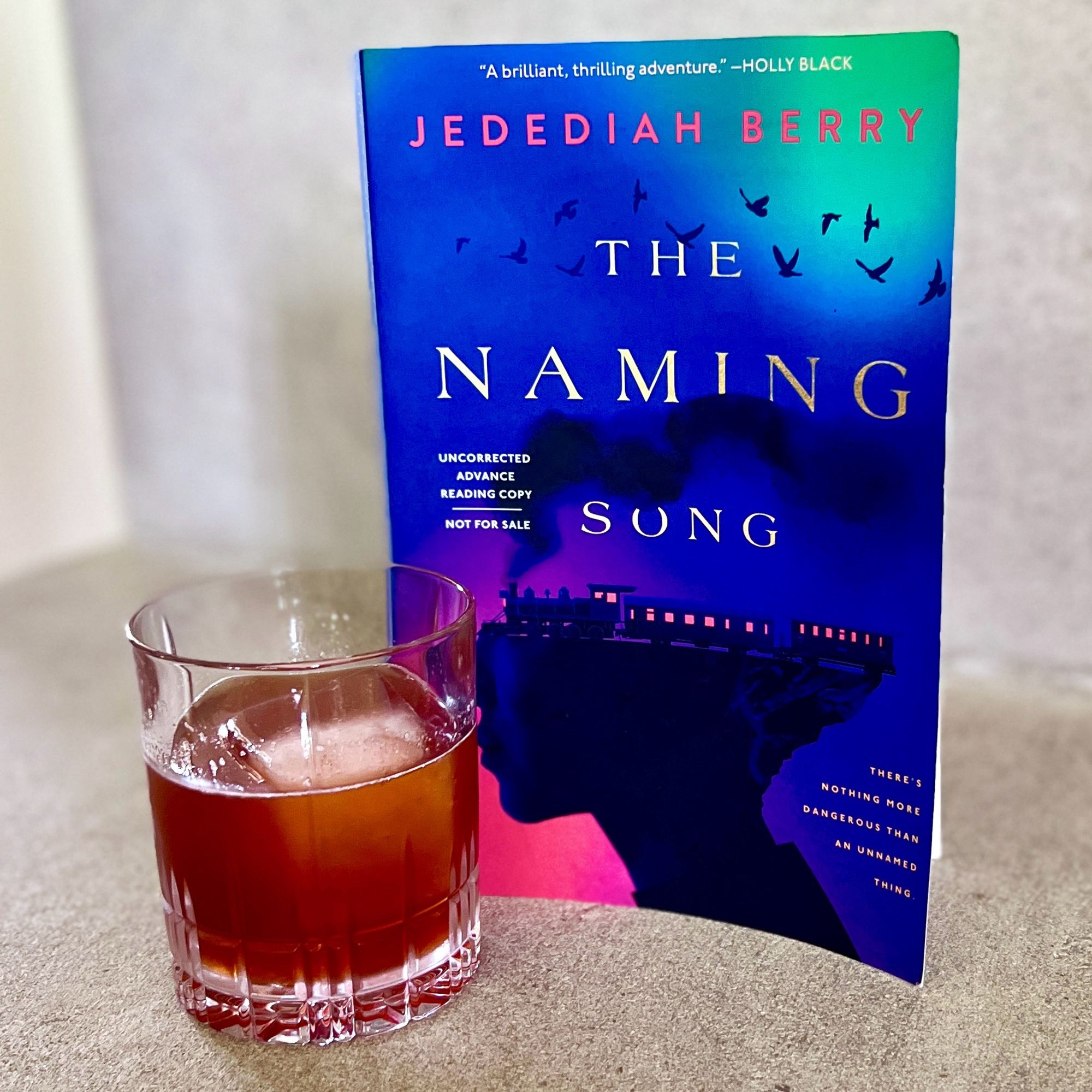 A dark reddish brown cocktail in an old fashioned glass alongside THE NAMING SONG by Jedediah Berry.