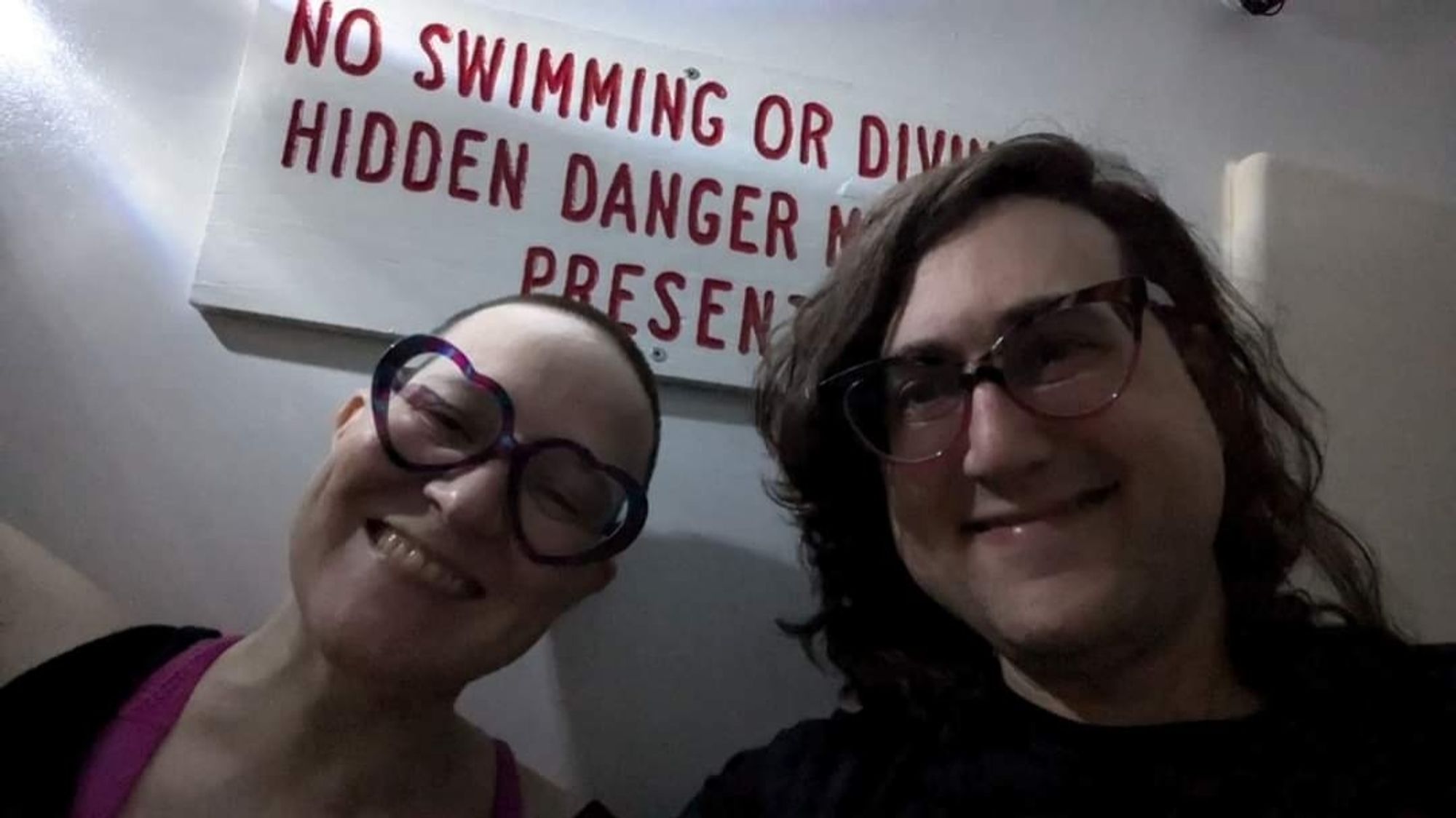 Two smiling white queerdos lit by a flashlight. The person in the left has their hair buzzed off and large heart shaped glasses, the person on the right has messy asymmetrical brown hair that's shoulder length at the longest and pink and shell cateye glasses. They're in front of a sign that reads NO SWIMMING OR DIVING HIDDEN DANGER MAYBE PRESENT.