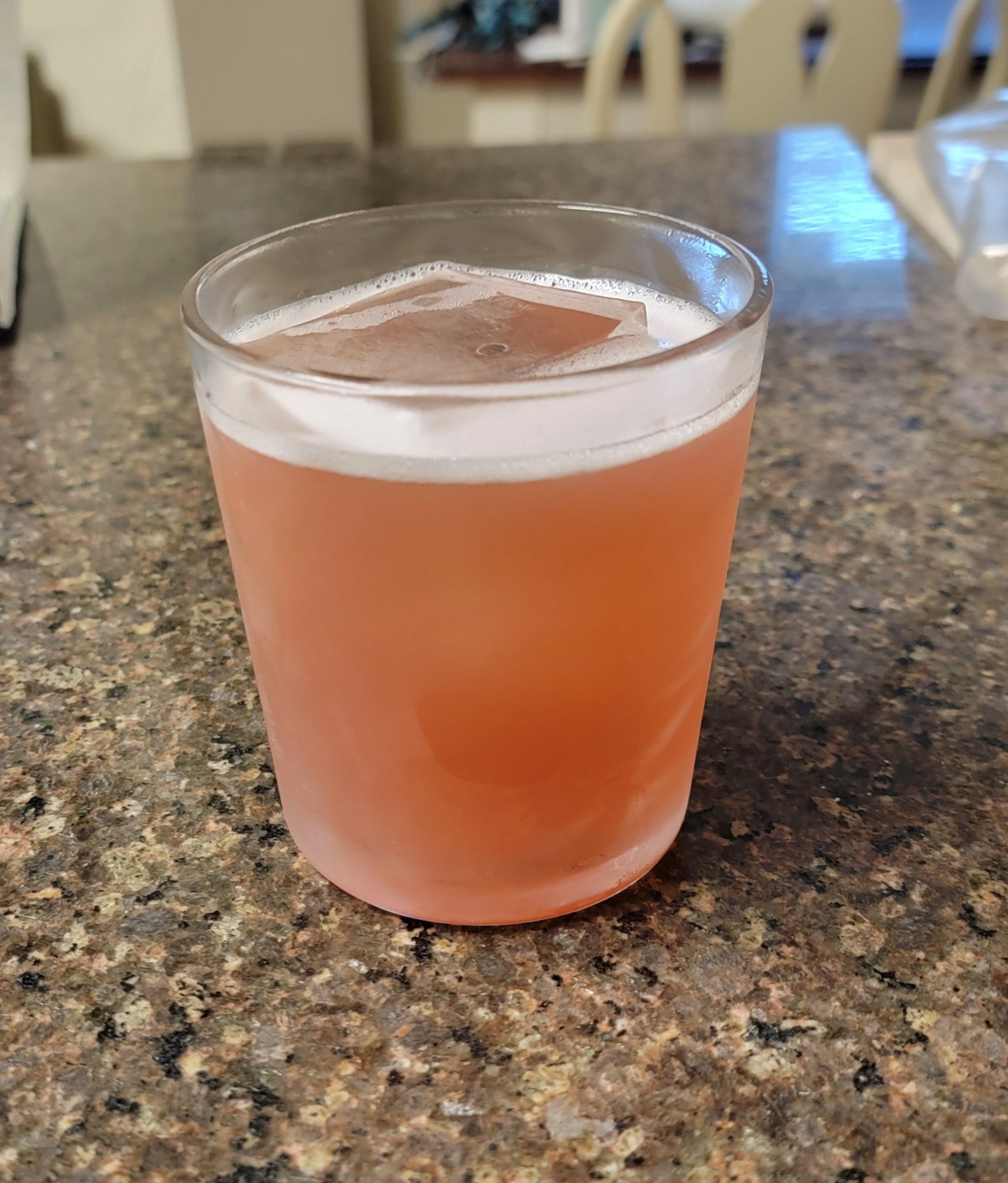 Redish-orange colored beverage in a glass. The glass is frosty and has a large ice cube in it