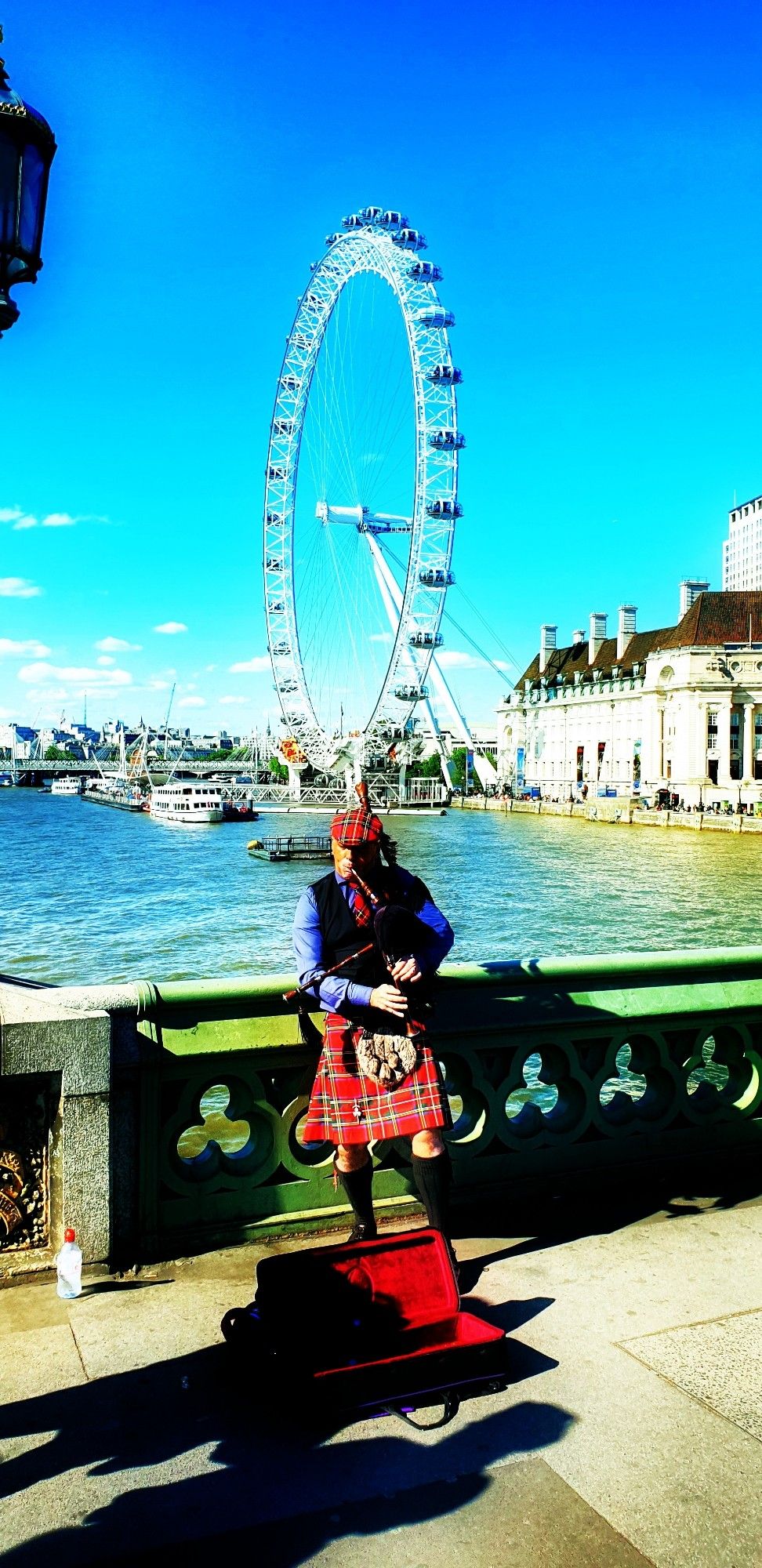 Das Bild zeigt eine Person, die einen traditionellen schottischen Kilt trägt und Dudelsack spielt. Im Hintergrund ist das London Eye, ein großes Riesenrad, zu sehen. Der Himmel ist blau mit einigen Wolken. Links im Bild fließt die Themse, und entlang des Flussufers sind Gebäude zu erkennen. Diese Szene spielt sich in London ab und bietet einen Blick auf eines der bekanntesten Wahrzeichen der Stadt.