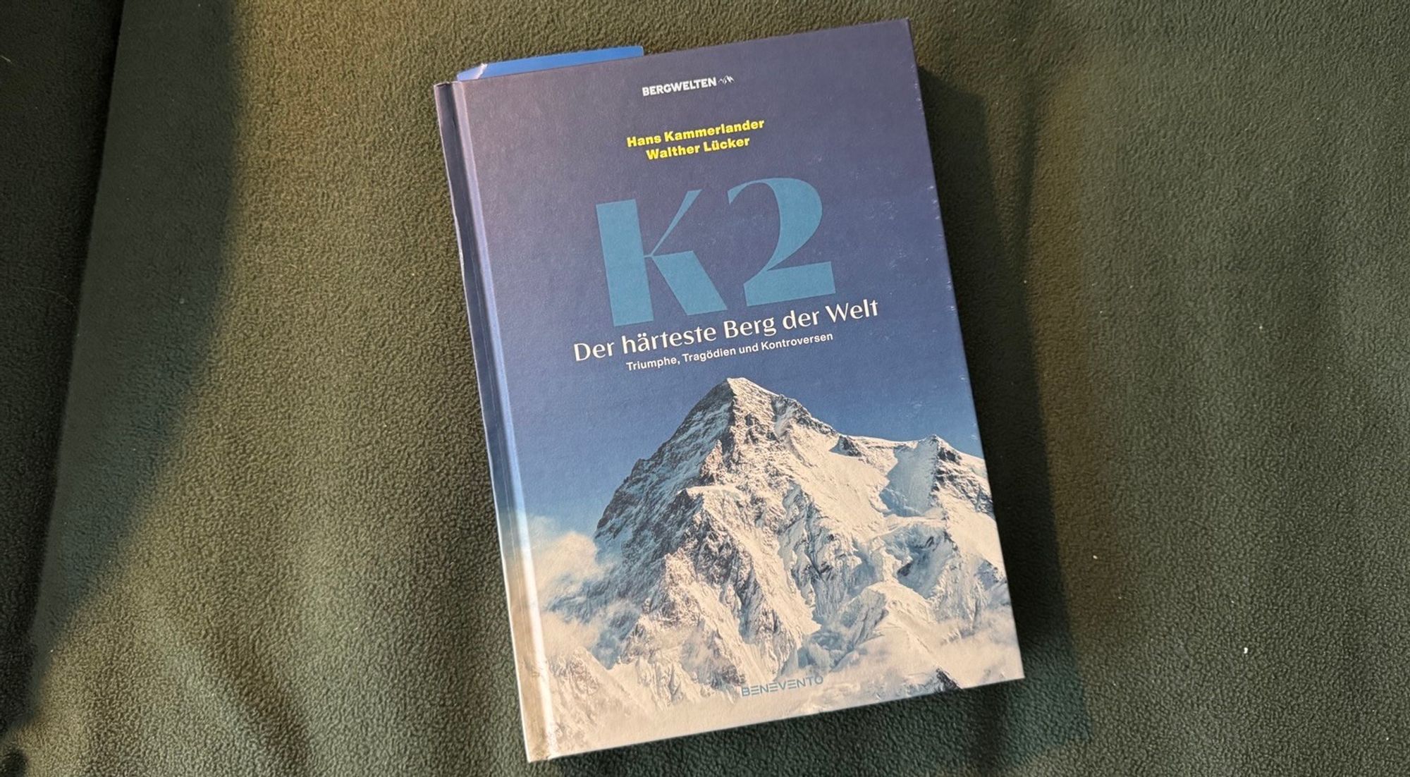 A book titled "K2: Der härteste Berg der Welt" by Hans Kammerlander and Walther Lücker, featuring a cover image of K2, the iconic mountain, with snow and rocky terrain. The background is a textured green