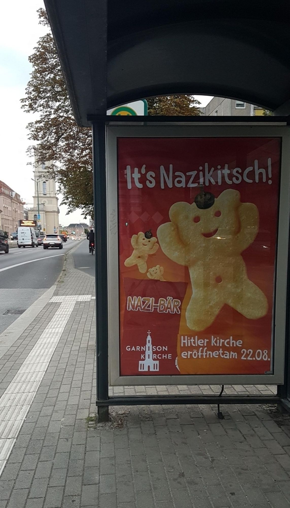 3x das gleiche Plakat an verschiedenen Haltestellen
roter/oranger Hintergrund
Überschrift: It's Nazikitsch!
dann ein großer und ein kleiner Pommbär  (Kartoffelchips) mit Pickelhaube und daneben: Nazi-Bär
unten:
links das neue GK-Turm Logo und der Abbinder: Hitler Kirche eröffnet am 22.08.