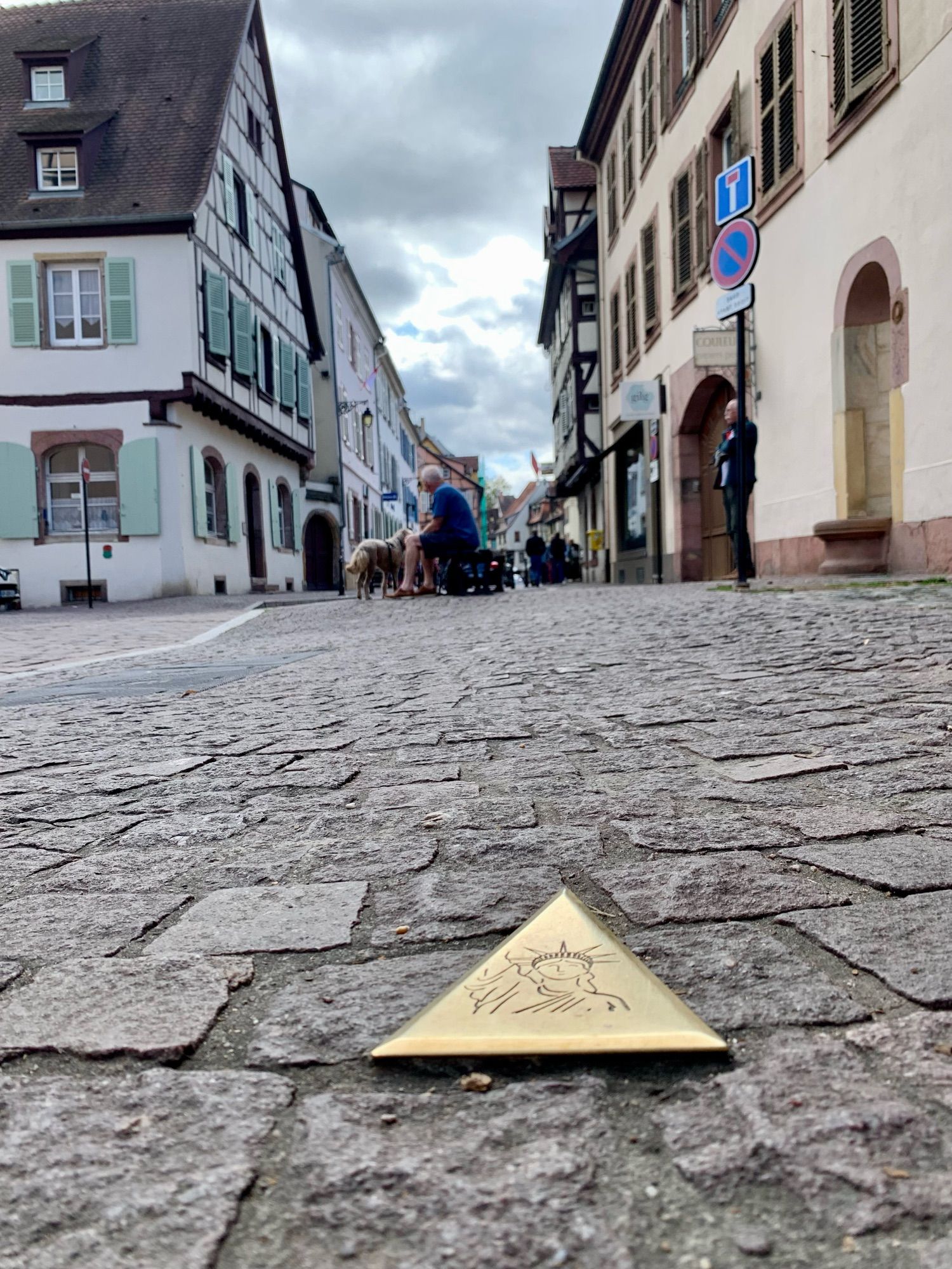 Straßenszene in Colmar, im Vordergrund ein im Pflaster eingelassenes Messingdreieck mit der Freiheitsstatue, im Hintergrund ein Mann mit Hund auf einer Bank.