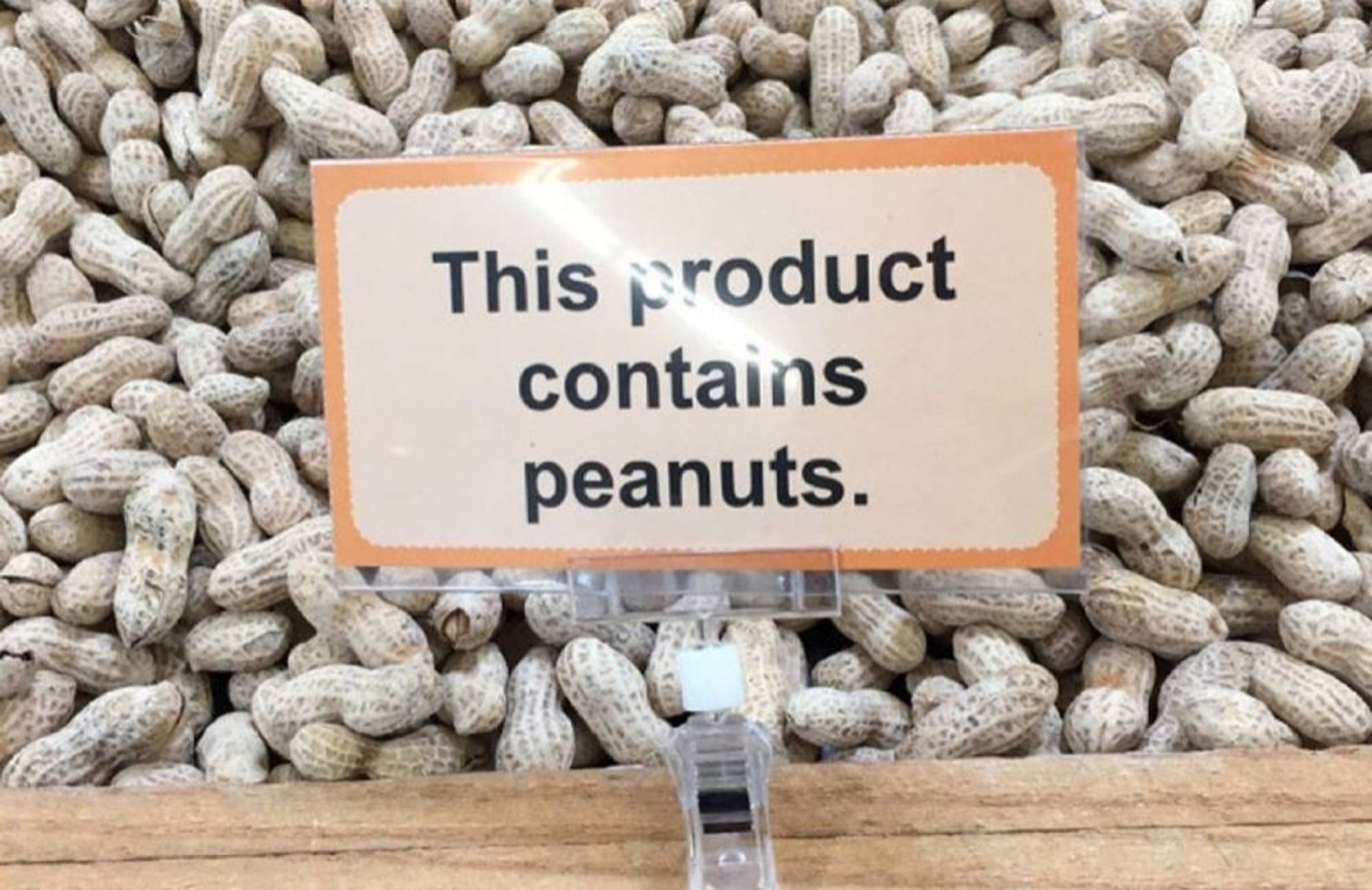 Sign in a shop saying "This product contains peanuts." It is in front if a clear-fronted container of unshelled peanuts